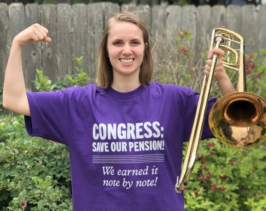 A young woman wearing a purple T-shirt that reads 'CONGRESS: SAVE OUR PENSION! We earned it note by note!' is standing outside in front of a wooden fence and plants, smiling and holding a brass trombone, with her other arm raised in a flexed muscle p