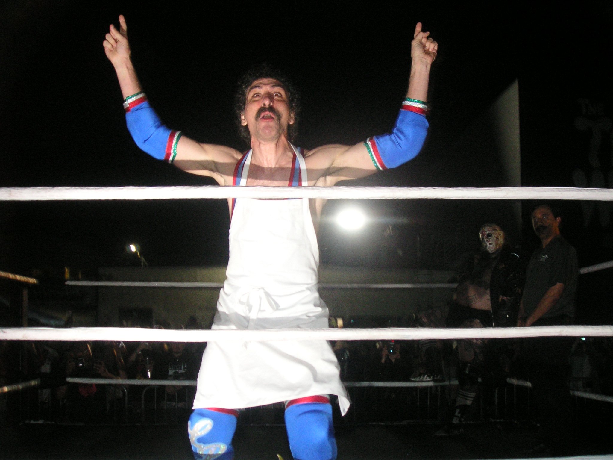 A man with a mustache and curly hair standing inside a boxing ring, wearing colorful boxing gear, with his arms raised and fingers crossed. Two people dressed as skeletons are in the background, one on the right and one on the left, partially visible