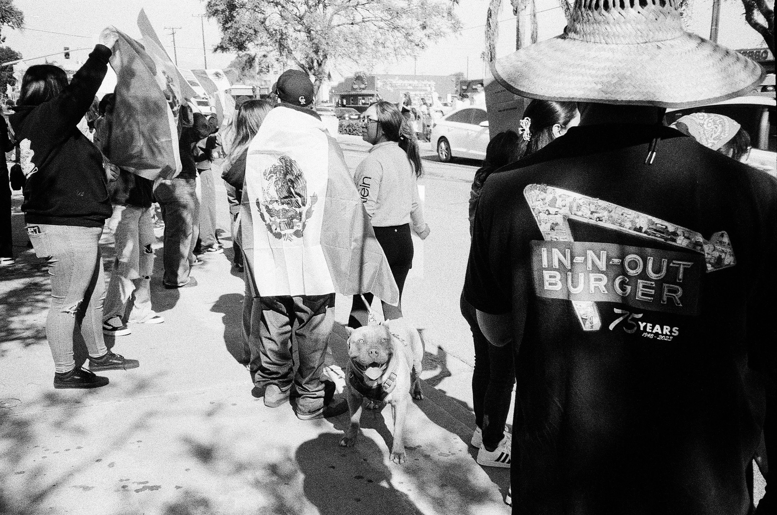 A group of people standing outdoors on a sidewalk, some holding flags and wearing casual clothing. A person in the foreground wears a jacket with 'In-N-Out Burger' logo, and a person nearby carries a flag with a graphic design, possibly a national em