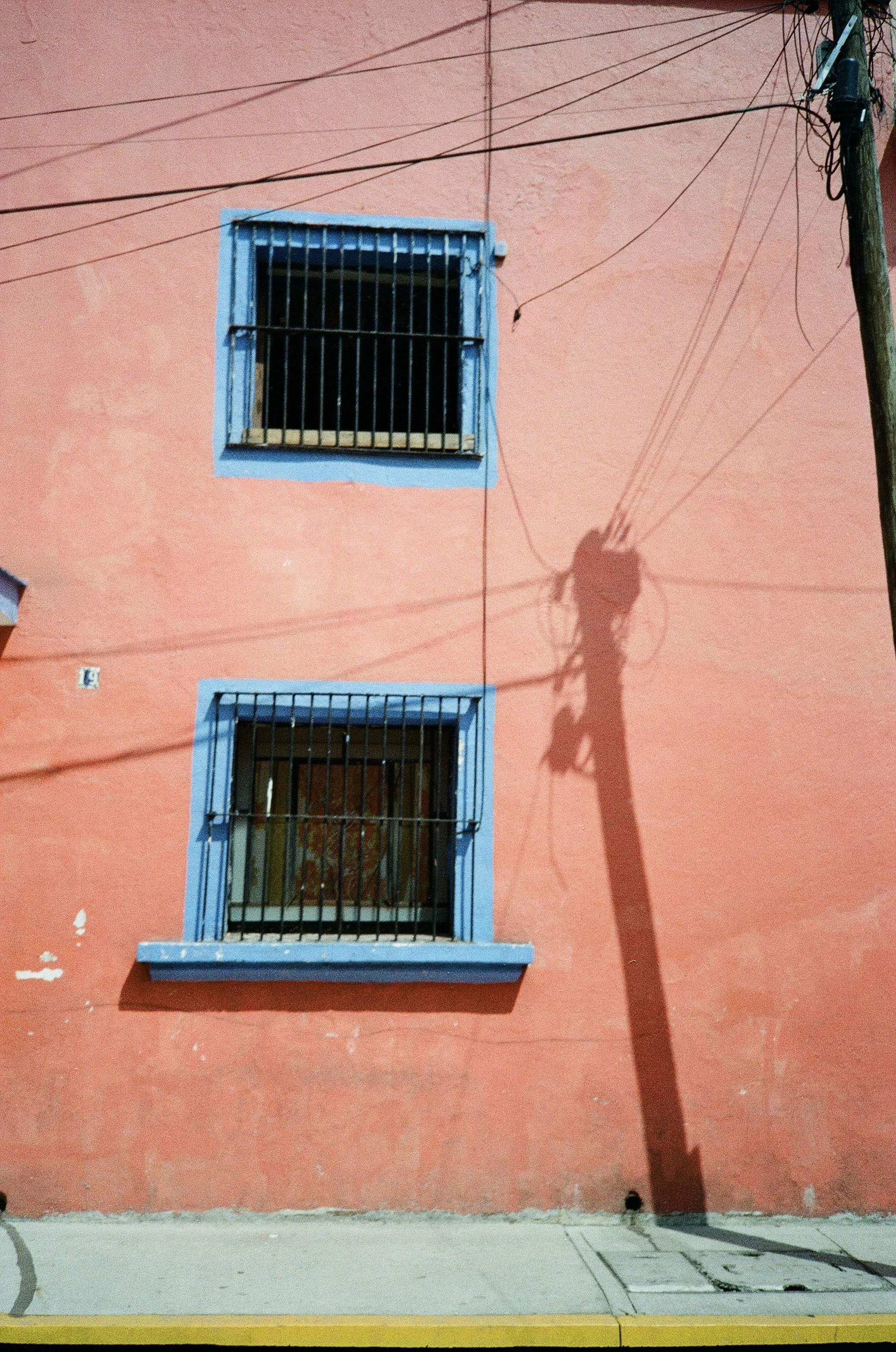 A pink building with three windows, each with blue frames and black metal bars, with a shadow of a utility pole and wires cast on the wall.