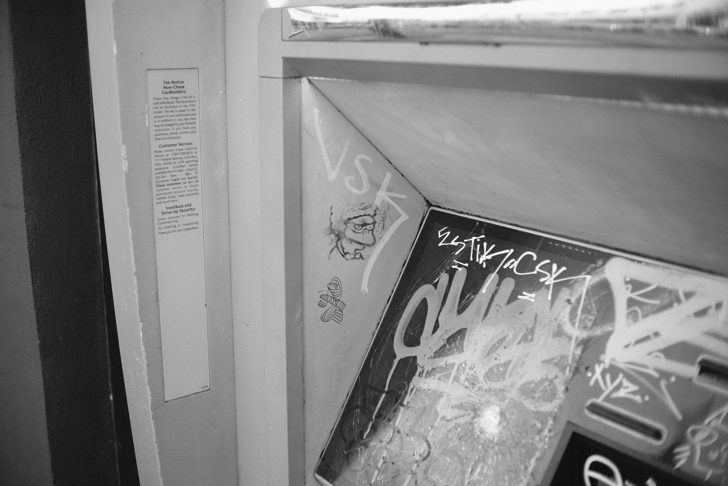 A black-and-white photograph of a section of an ATM machine, featuring graffiti writing and illustrations on its surface, with a white fee notice sticker on the adjacent wall.