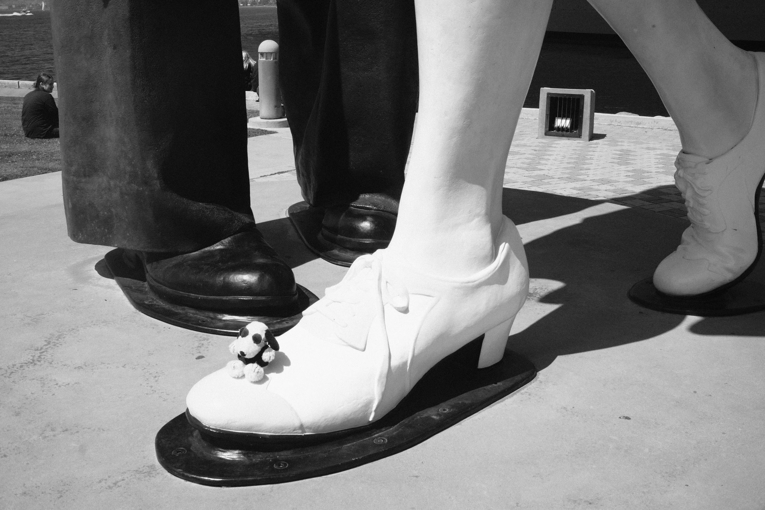 Close-up of a sculpture of shoes, with a small stuffed dog toy placed on the sculpture. In the background, people are sitting near a body of water.