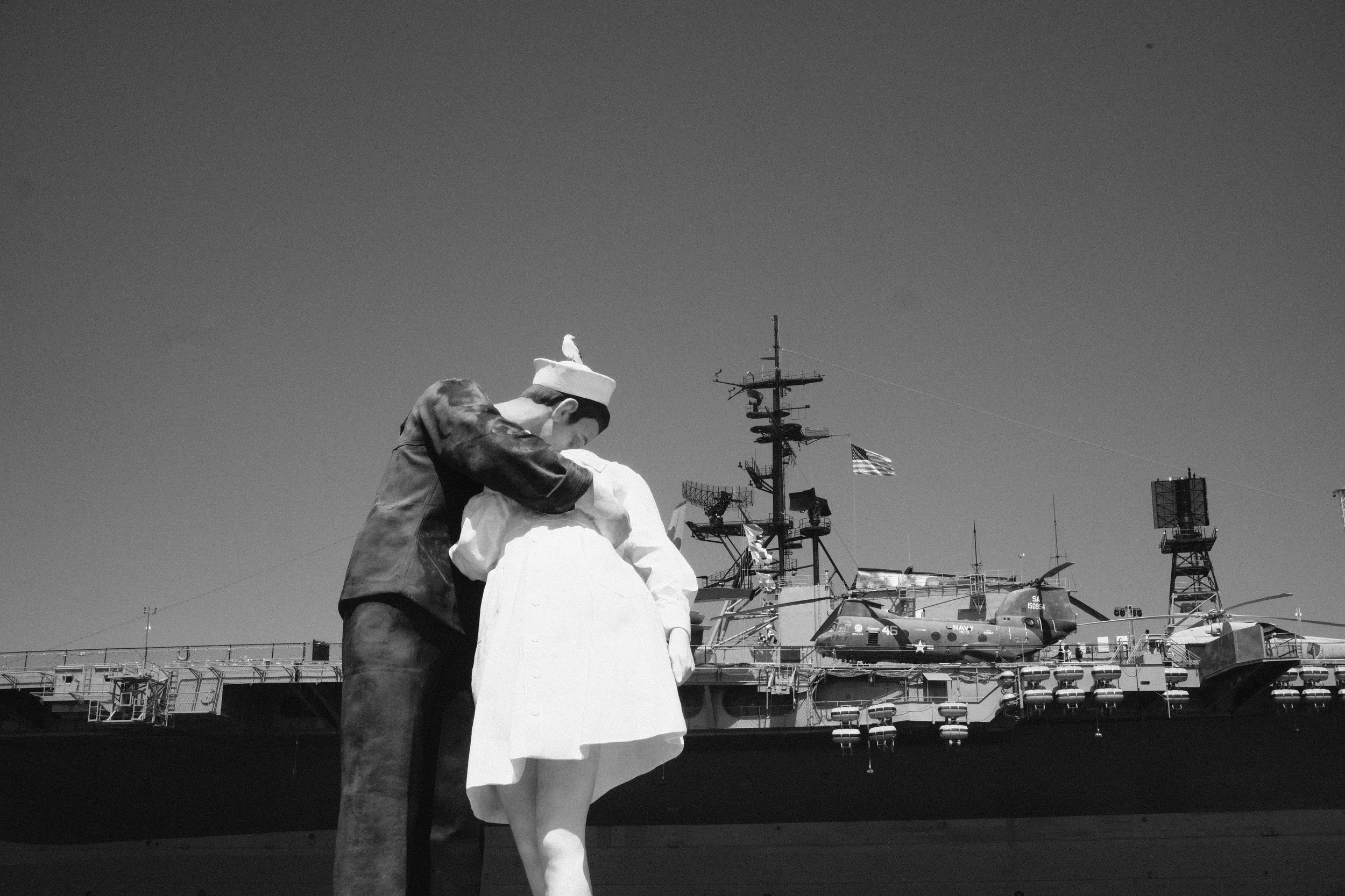 Black and white photo of two sailors embracing on the deck of a naval ship, with boats and radio antennas in the background, and an American flag flying.