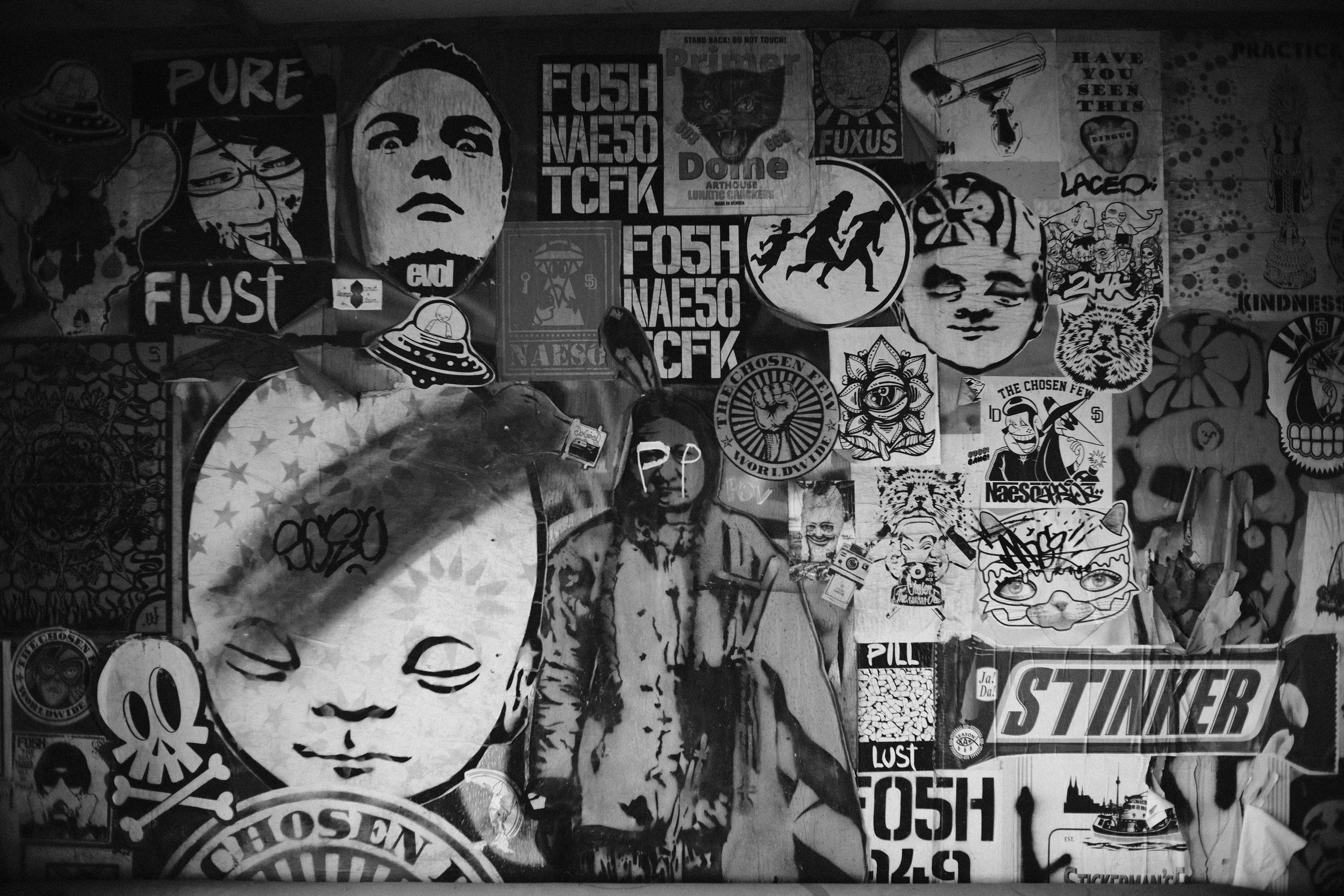 A collage of various black and white stickers and posters on a wall, including portraits, symbols, text, and illustrations.