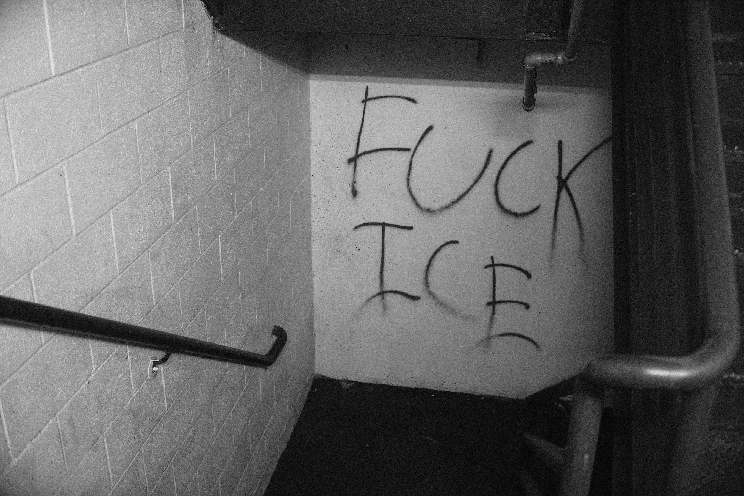 A black-and-white photo of graffiti on a wall near a pipe and a handrail. The graffiti includes the words "FUCK TOS" or similar.