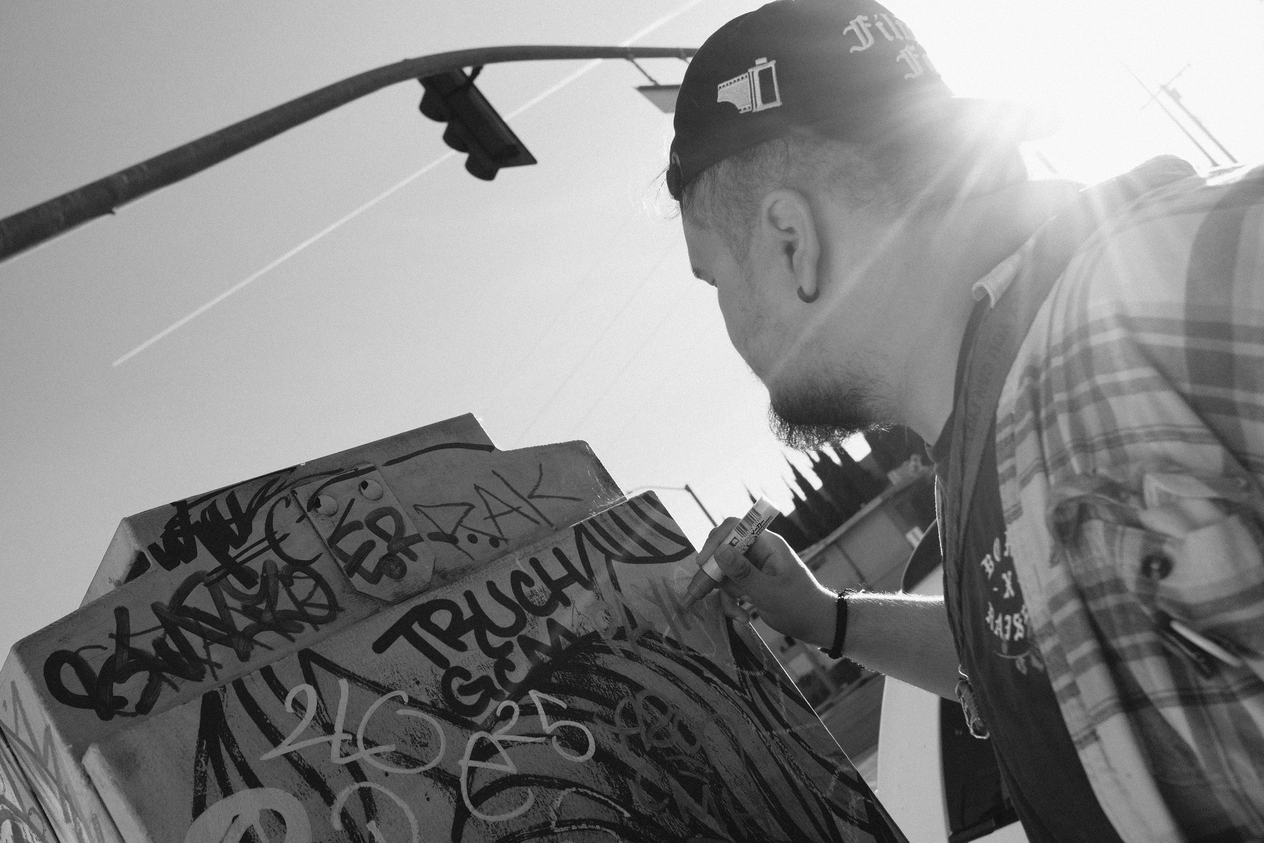 A person with short hair, a beard, earrings, and a bandana on their head is spray painting graffiti on a wall or large surface outdoors. The background shows a clear sky and utility poles.