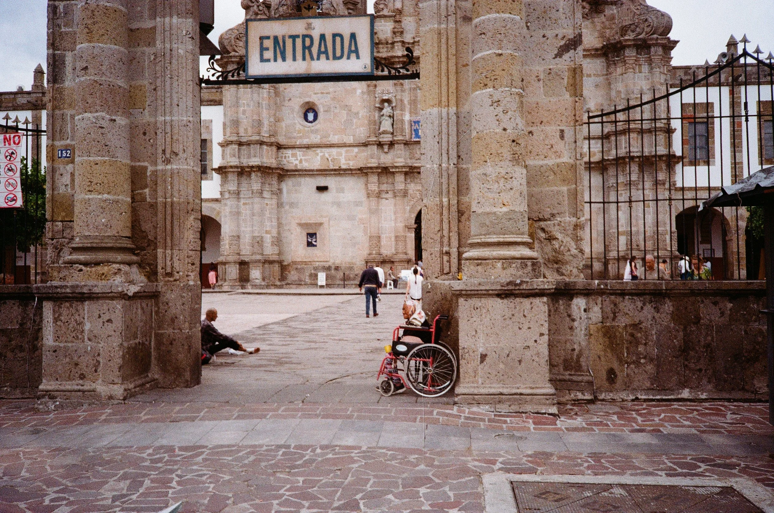 A view through an old stone archway of a historic building and plaza with people walking and sitting, and a wheelchair parked in the foreground.