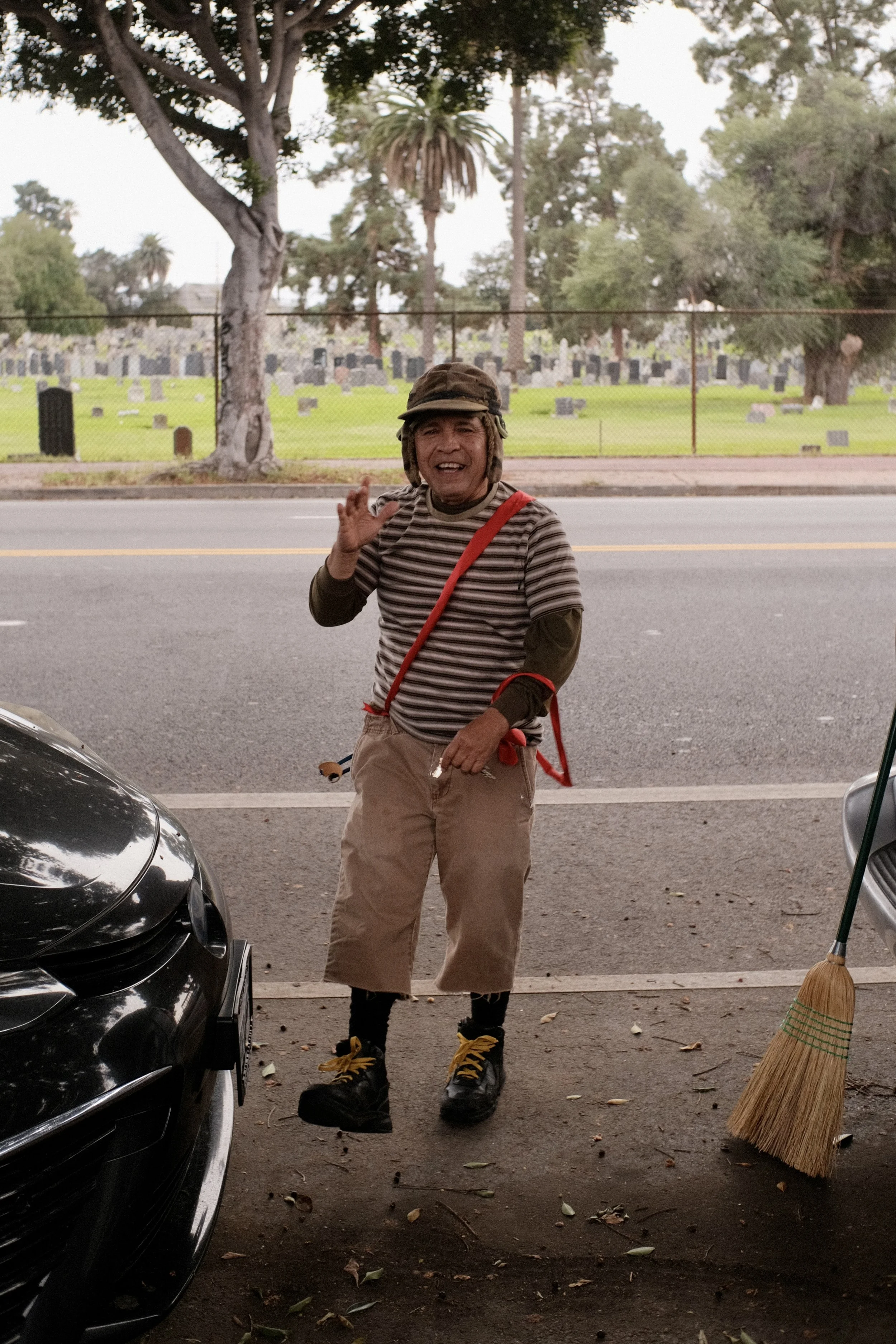 A person smiling and waving outdoors, standing near a black car on a street, wearing a striped shirt, tan pants, black boots, and a helmet. A fence and a cemetery with headstones and trees are visible in the background, along with a broom and a leaf 