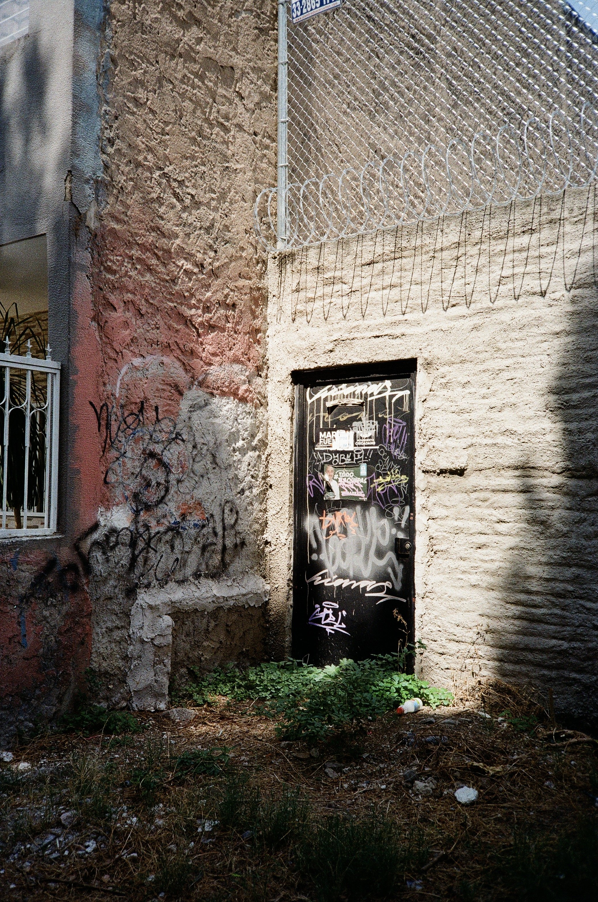A small black door on a weathered beige wall, covered with graffiti, with overgrown grass and trash at the base, and barbed wire above on a rusty chain-link fence.