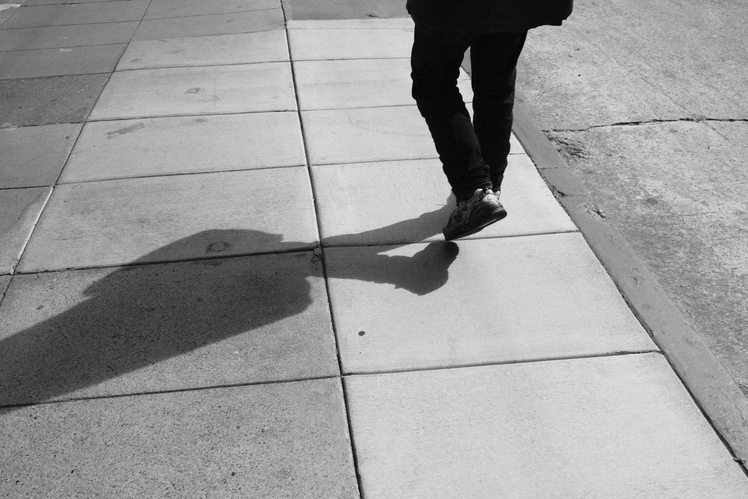 A person walking on a sidewalk, dressed in black pants and shoes, with their shadow visible on the pavement.