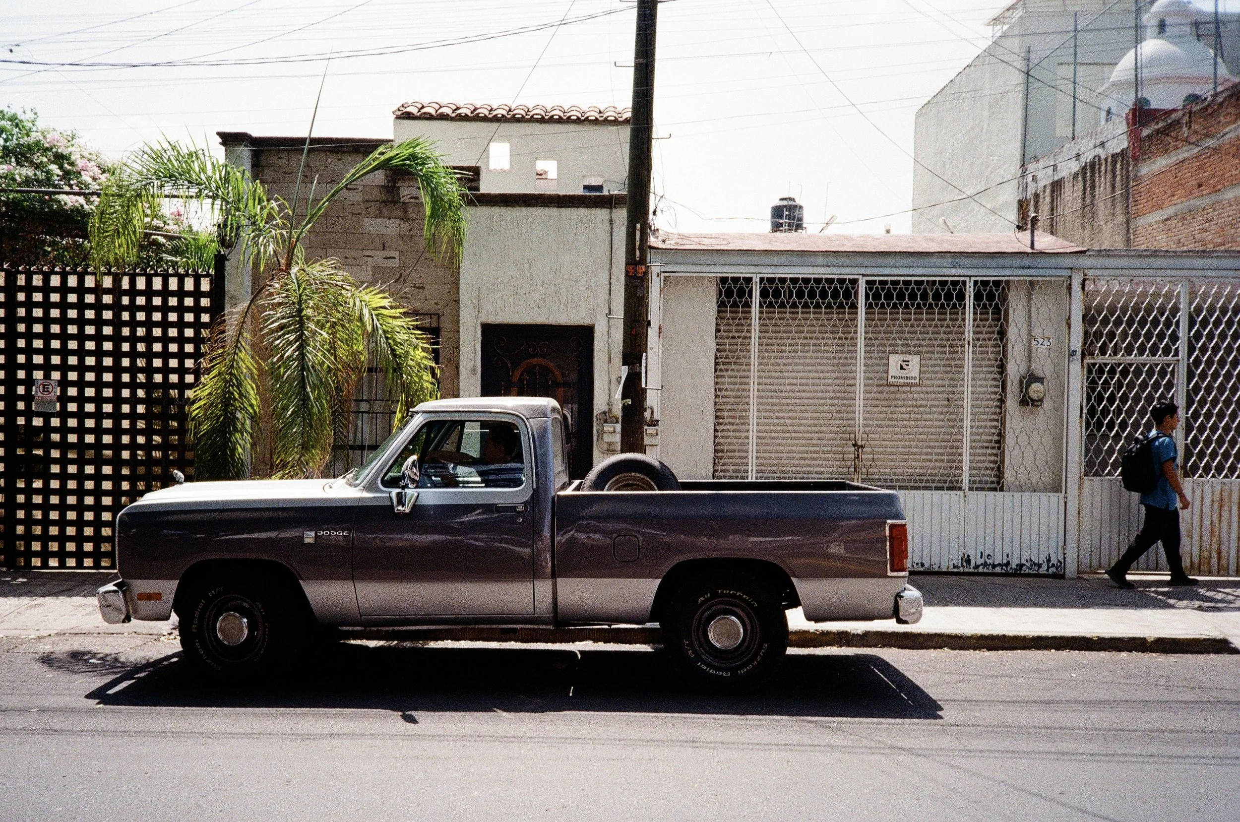 A black pickup truck driving on a city street with a person walking on the sidewalk and residential buildings in the background.