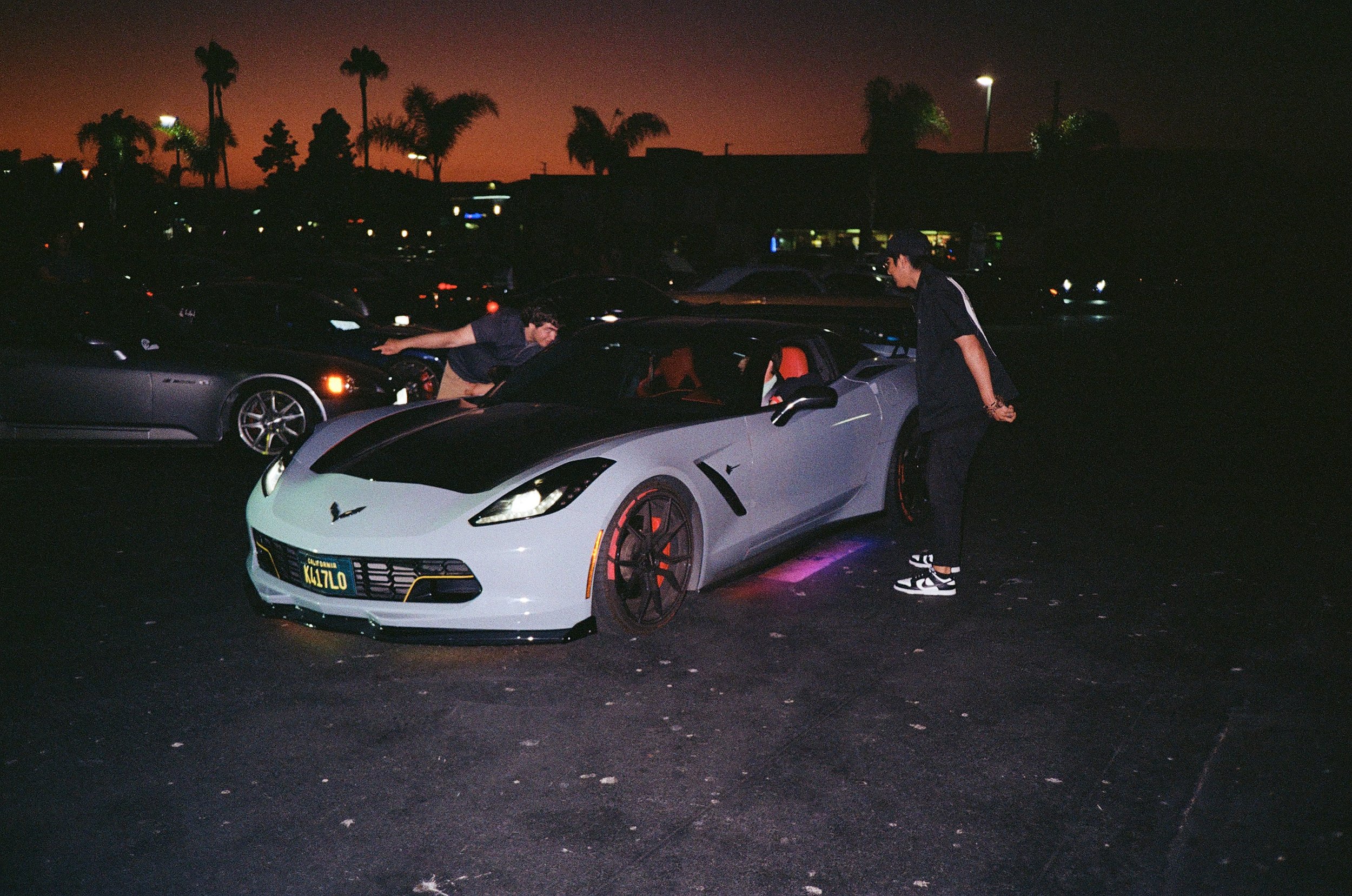 Nighttime scene in a parking lot with two young men, one leaning over a gray sports car and the other standing beside it, with palm trees and a darkening sky in the background.