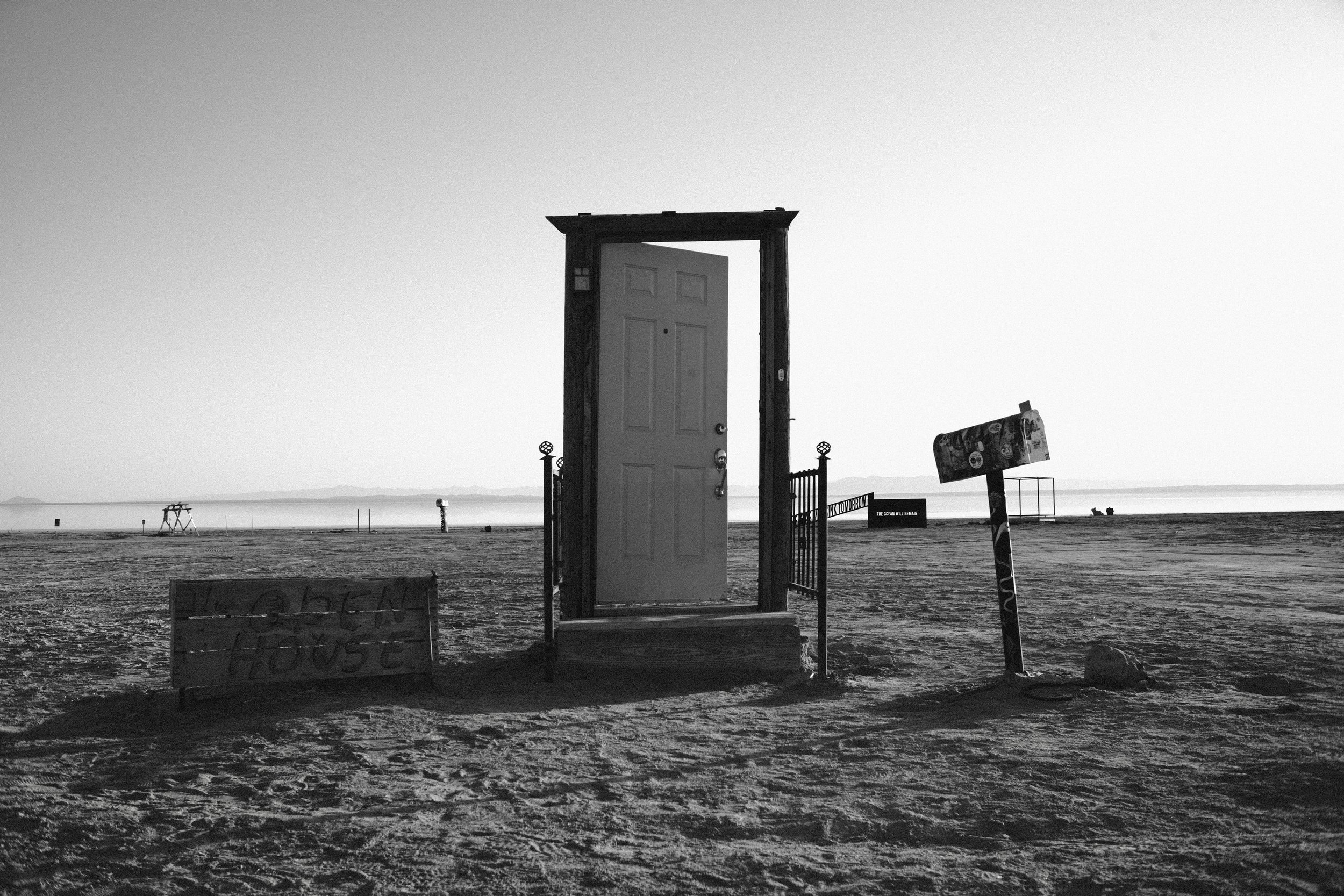Empty door standing alone in the middle of a plain landscape, with a small sign that reads "OPEN HOUSE" in front of it. The scene is in black and white, with a clear sky and mountains in the background.