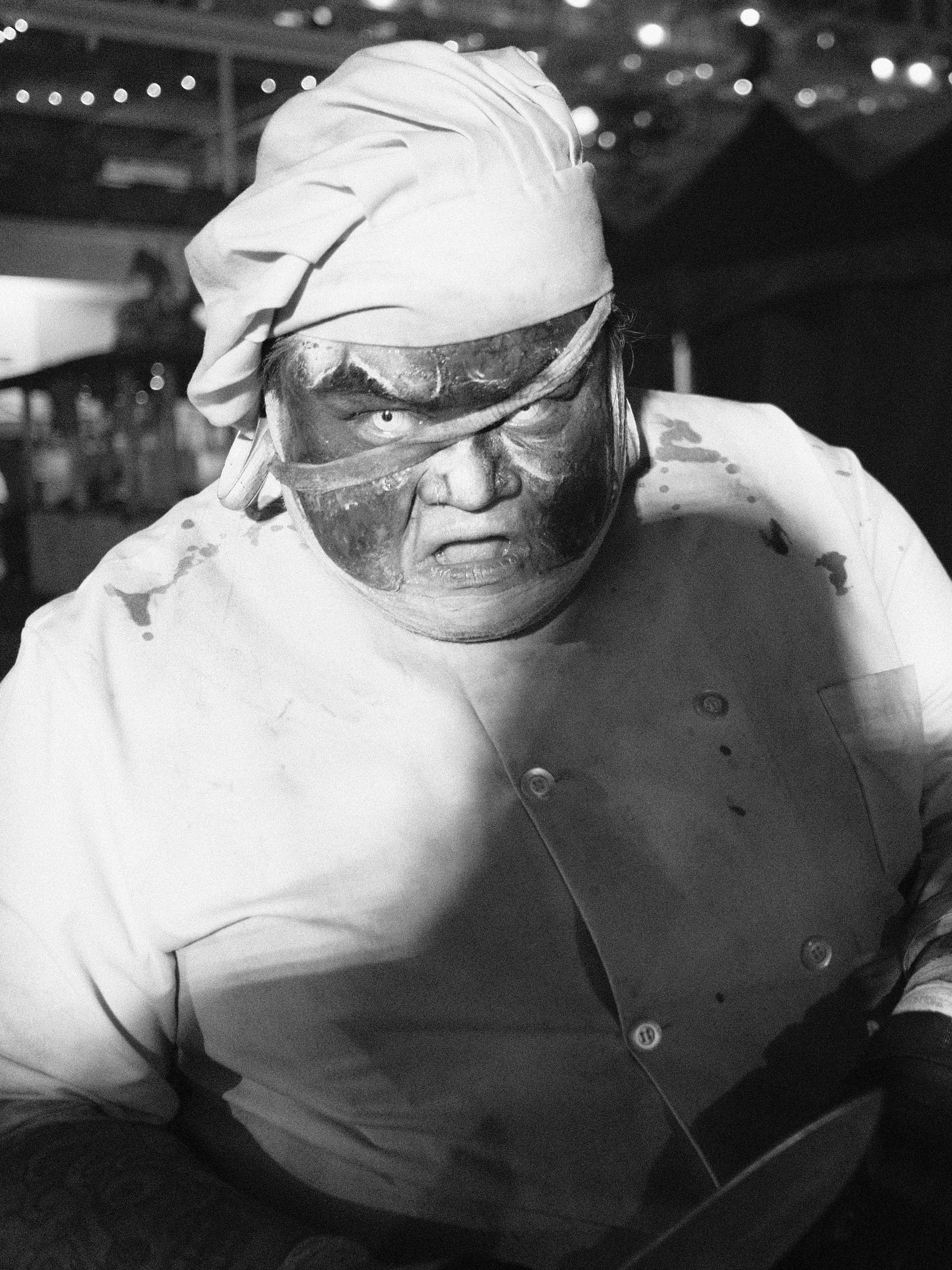 A chef wearing a white chef's coat and a head covering, with paint on his face and clothing, appears to be upset or angry in a kitchen or restaurant setting.