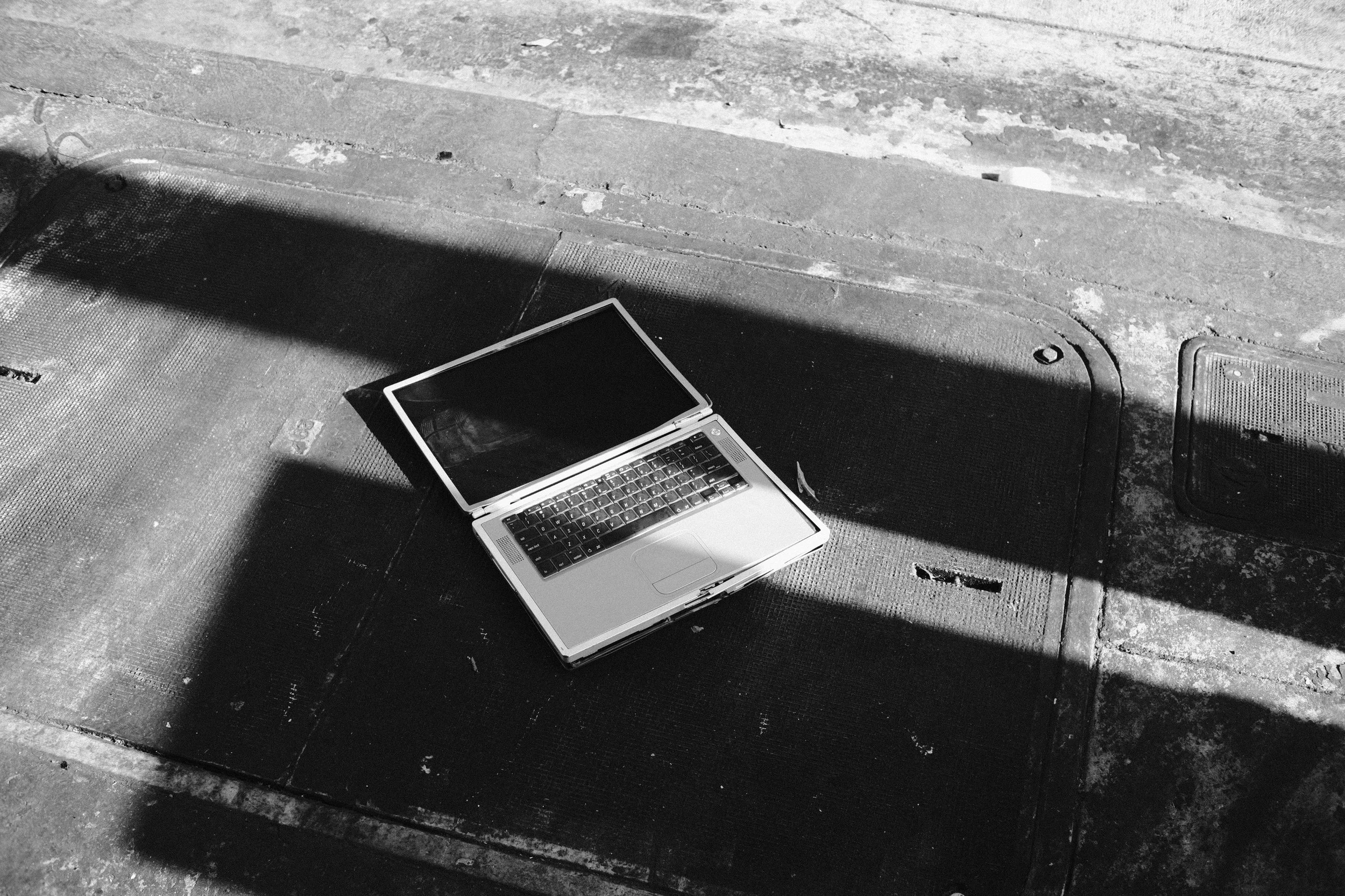 A laptop resting on an outdoor concrete surface with scattered debris, partially shaded by a shadow.