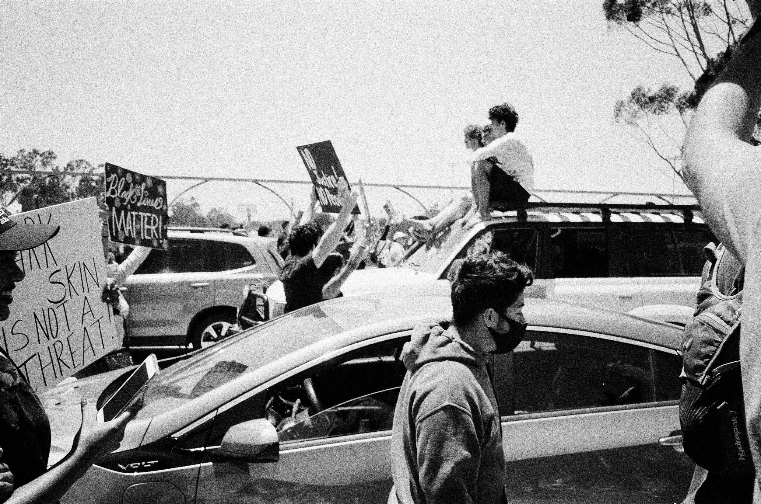 A black and white photo of a protest with people holding signs with messages about racial justice, including 'Black Lives Matter' and 'This is not a threat.' People are wearing masks and some are standing on or near cars.