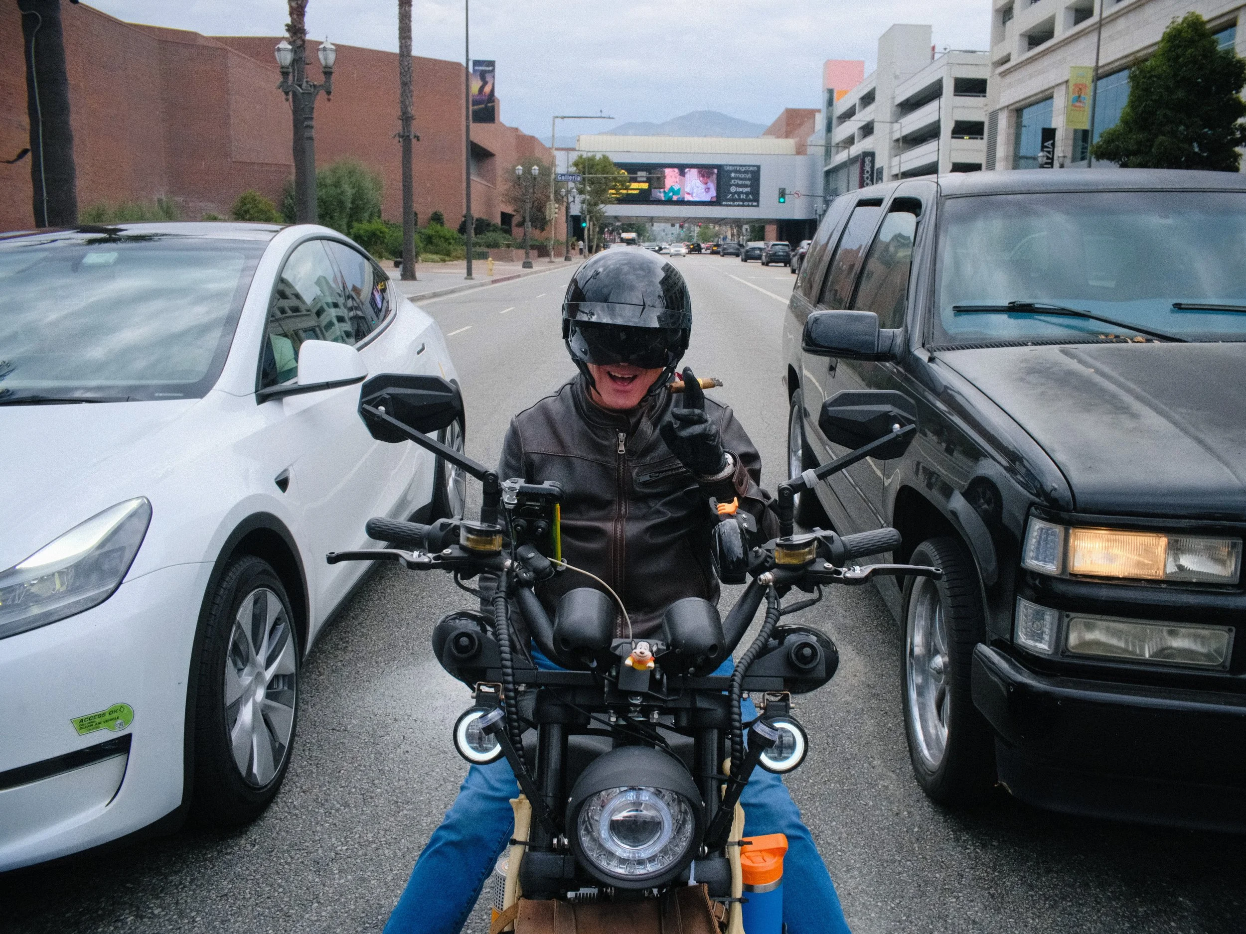 A person wearing a black helmet and leather jacket is sitting on a black motorcycle in a city street, smiling and giving a thumbs up. The street is lined with parked cars, and a digital billboard is visible in the background.