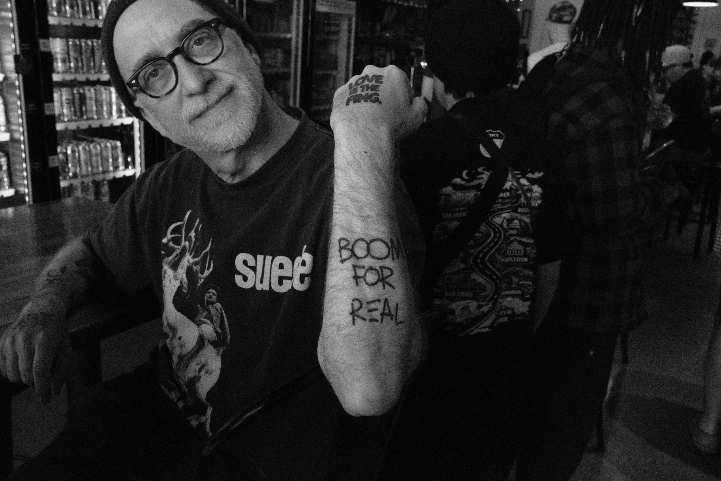 A man with a beard and glasses smiling at the camera, showing a tattoo on his right arm that reads "BOOM FOR REAL" and a tattoo on his hand that says "LOVE IS THE FING." He is sitting in a room with bookshelves and other people in the background, som