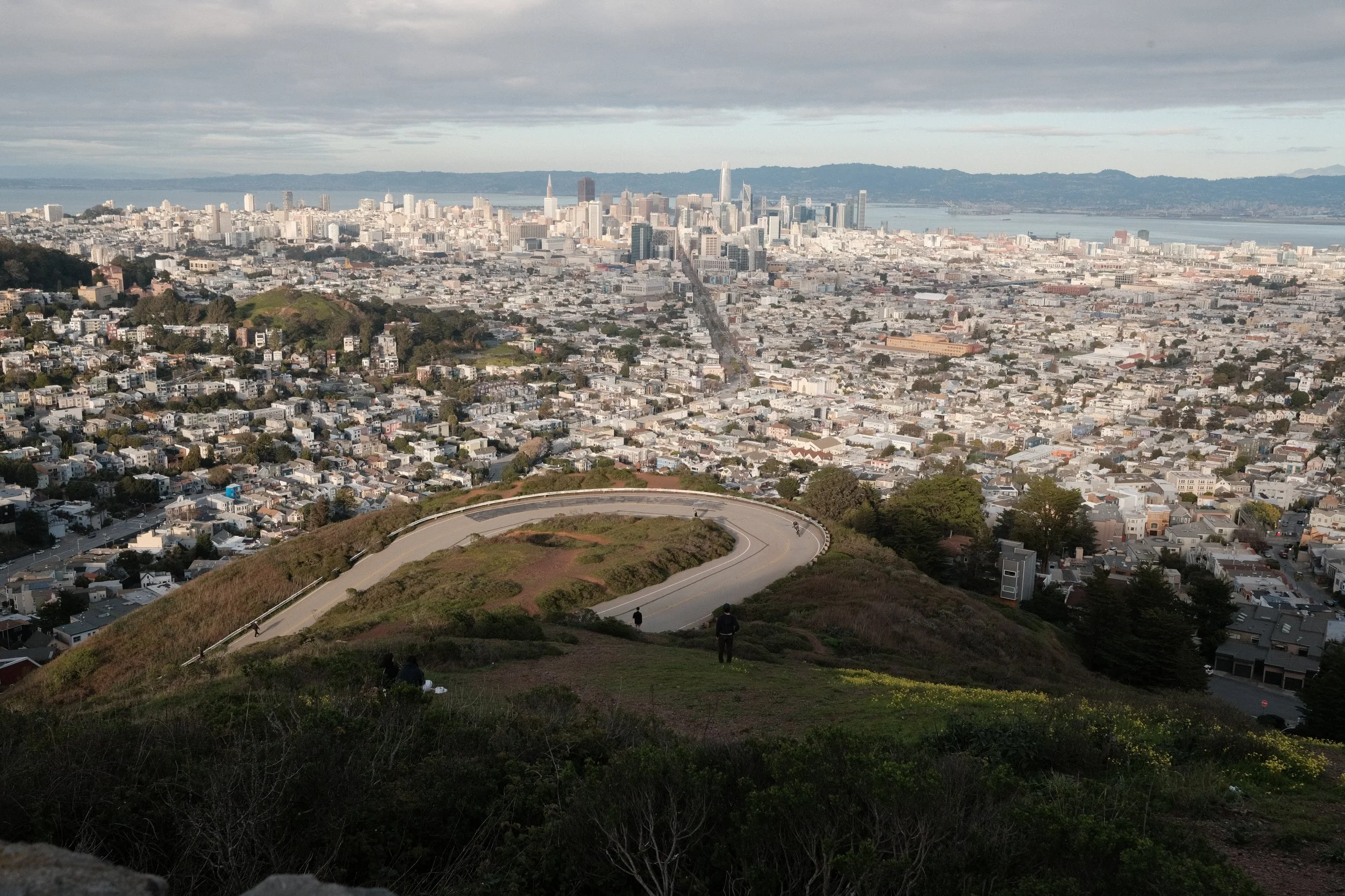 A panoramic view of San Francisco skyline, seen from a hill with a winding road and some people sitting and walking on the grassy area in the foreground.