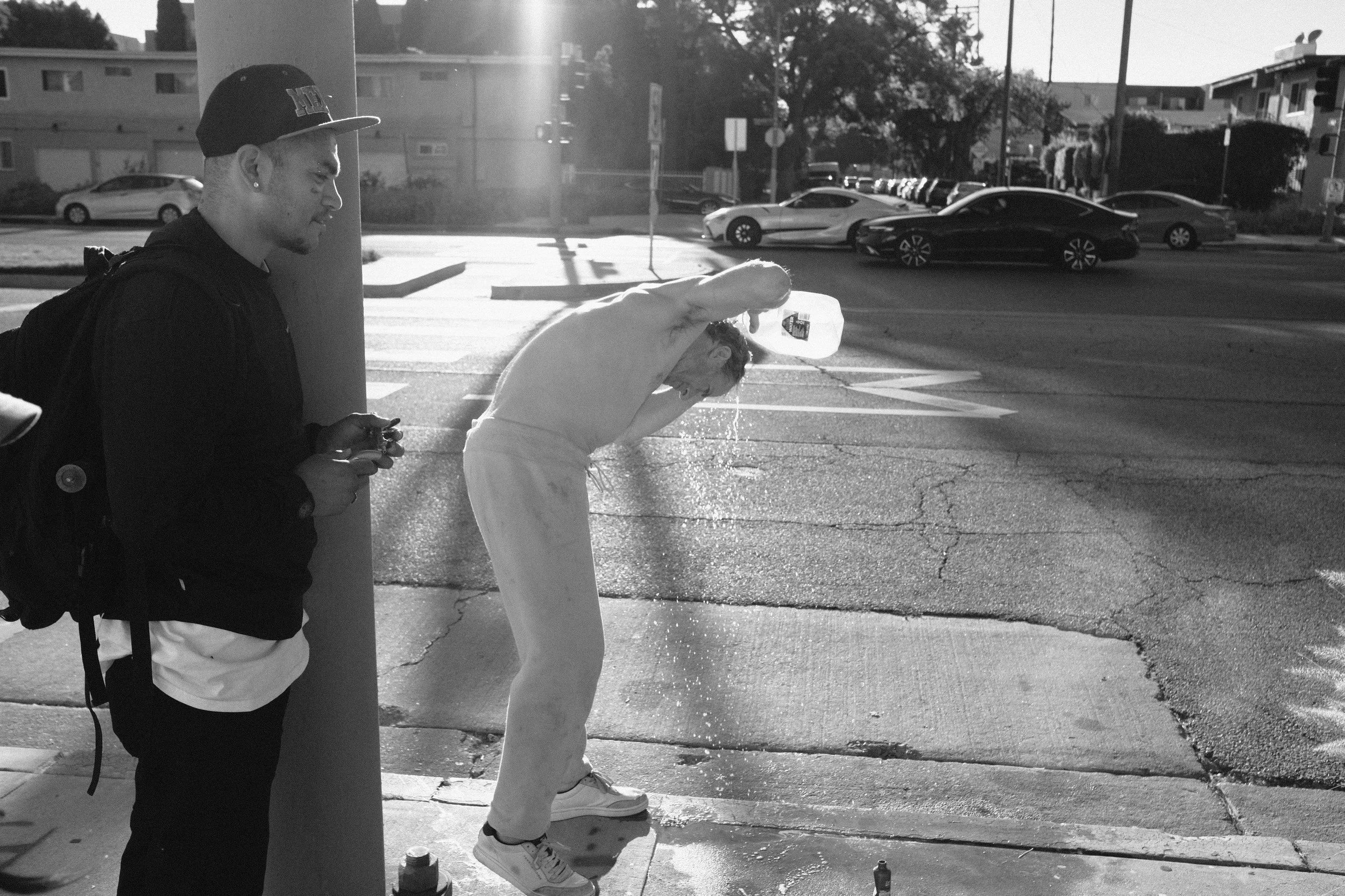 A black-and-white photo of a young man in a white hoodie and white sneakers skateboarding on a city sidewalk, with two men standing against a pole watching. Parked cars and residential buildings are visible in the background, with sunlight casting sh