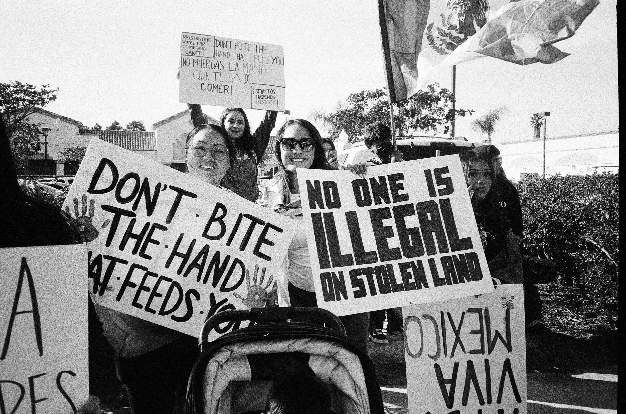 Group of women protesting outdoors, holding signs with messages in English and Spanish, including "No one is illegal" and "Don't bite the hand that feeds you," with trees and buildings in the background.