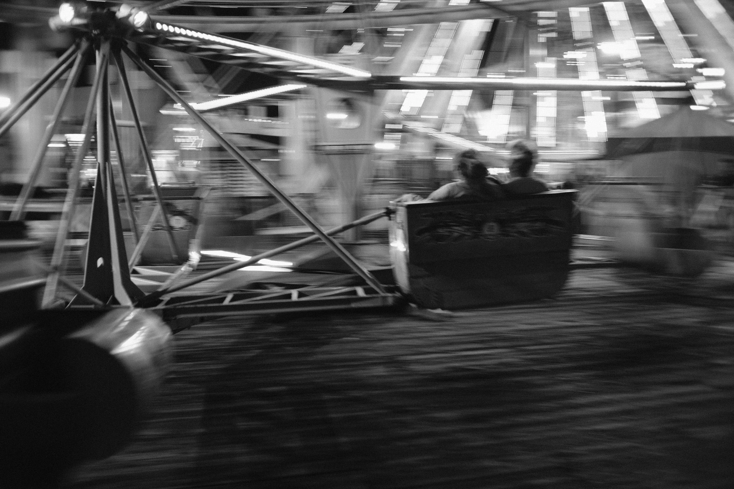 Black and white blurred photo of a carousel or amusement park ride with two people sitting on a bench, motion blur giving a sense of movement.