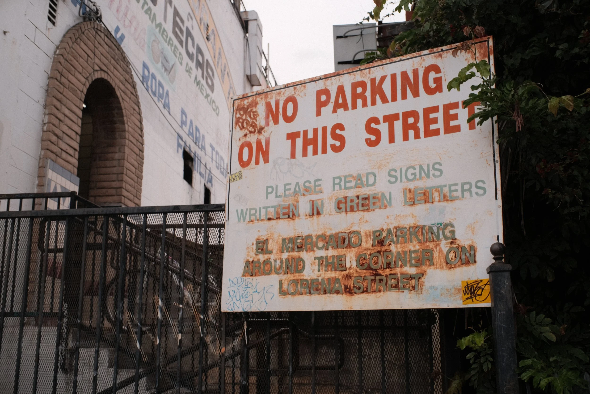 A weathered sign that says 'No Parking on This Street' and additional instructions written in green lettering about reading signs and written in green letters, mounted near a black metal fence and leafy green plants. A building with brick and white w