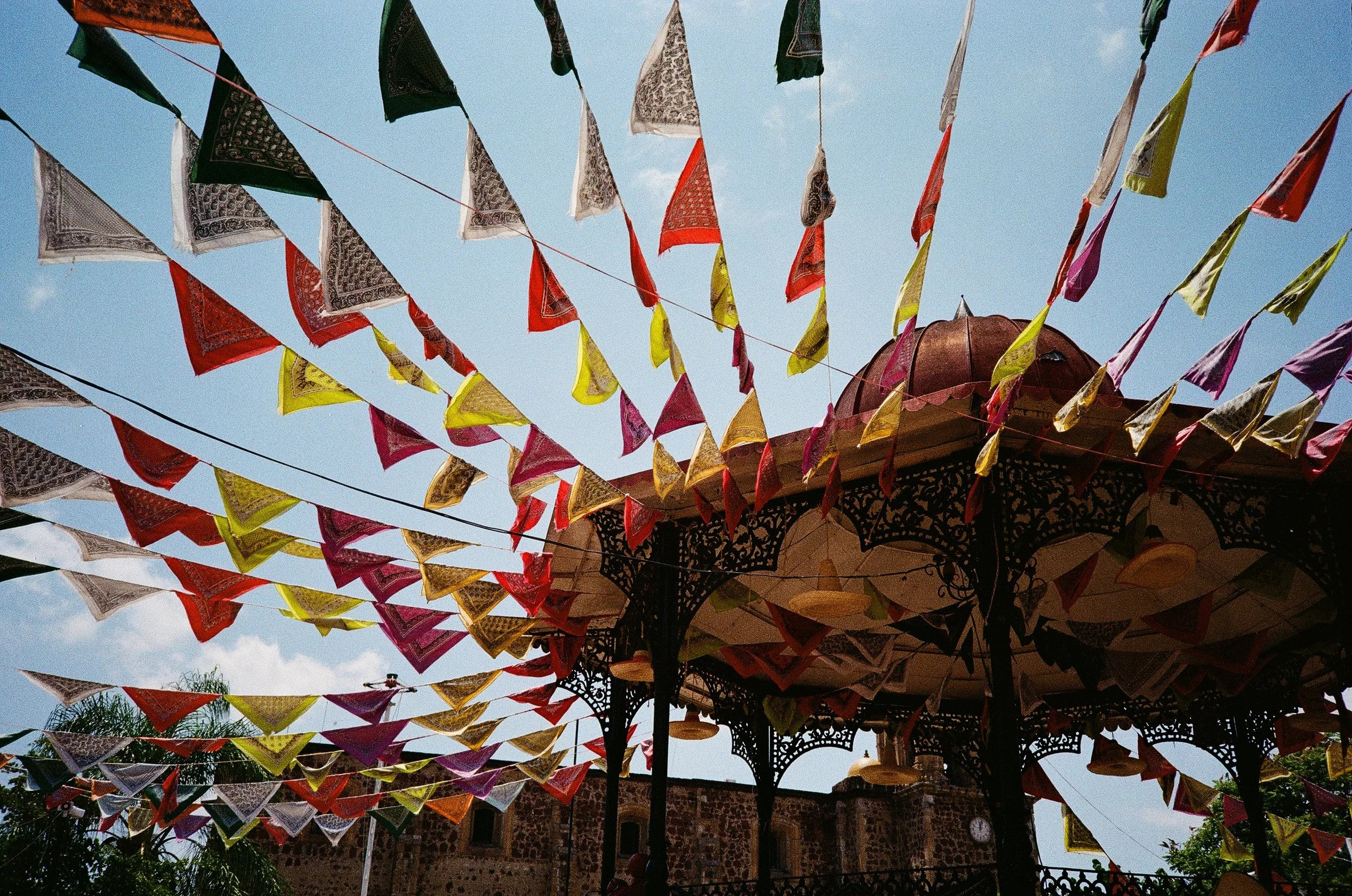 Colorful triangular fabric banners hanging from lines in an outdoor setting with a decorative structure visible beneath the banners, set against a blue sky.