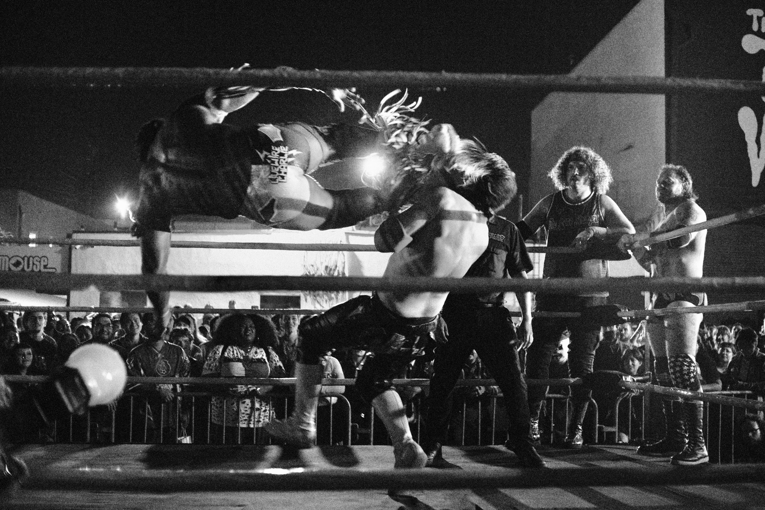 A black and white photo of a women's wrestling match taking place in front of a large crowd at night, with several women grappling inside a wrestling ring and spectators watching around the ring.