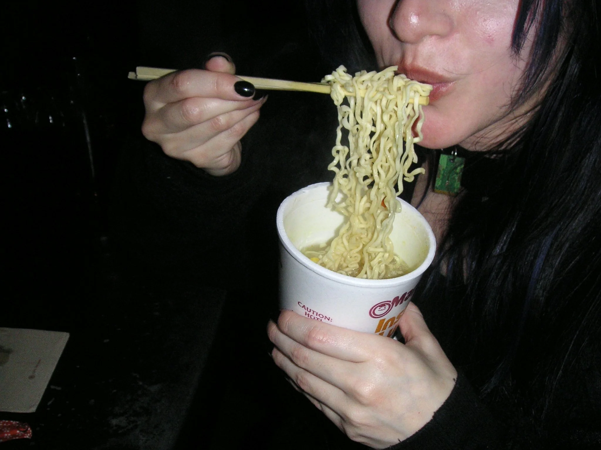 A person with dark hair eating noodles with chopsticks, holding a cup of instant noodles in their left hand.