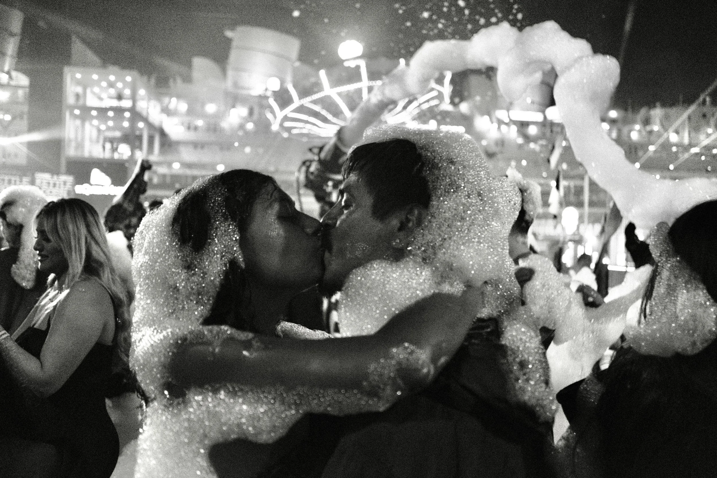 Two people are embracing and about to kiss at a foam party, with foam covering their bodies and faces, at a crowded venue with bright lights in the background.
