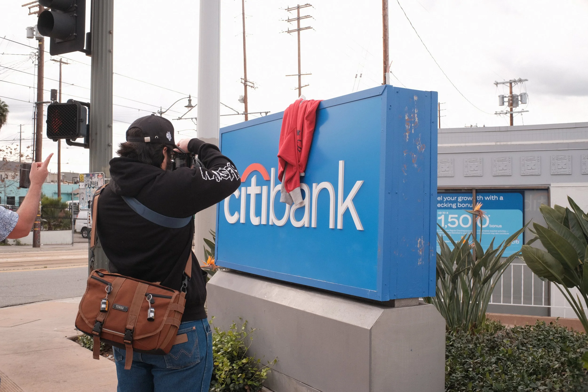 A person taking a photograph of a large blue sign with the text 'Citzbank' on it, in an outdoor urban setting with power lines and buildings in the background, and a red shirt draped over the top of the sign.