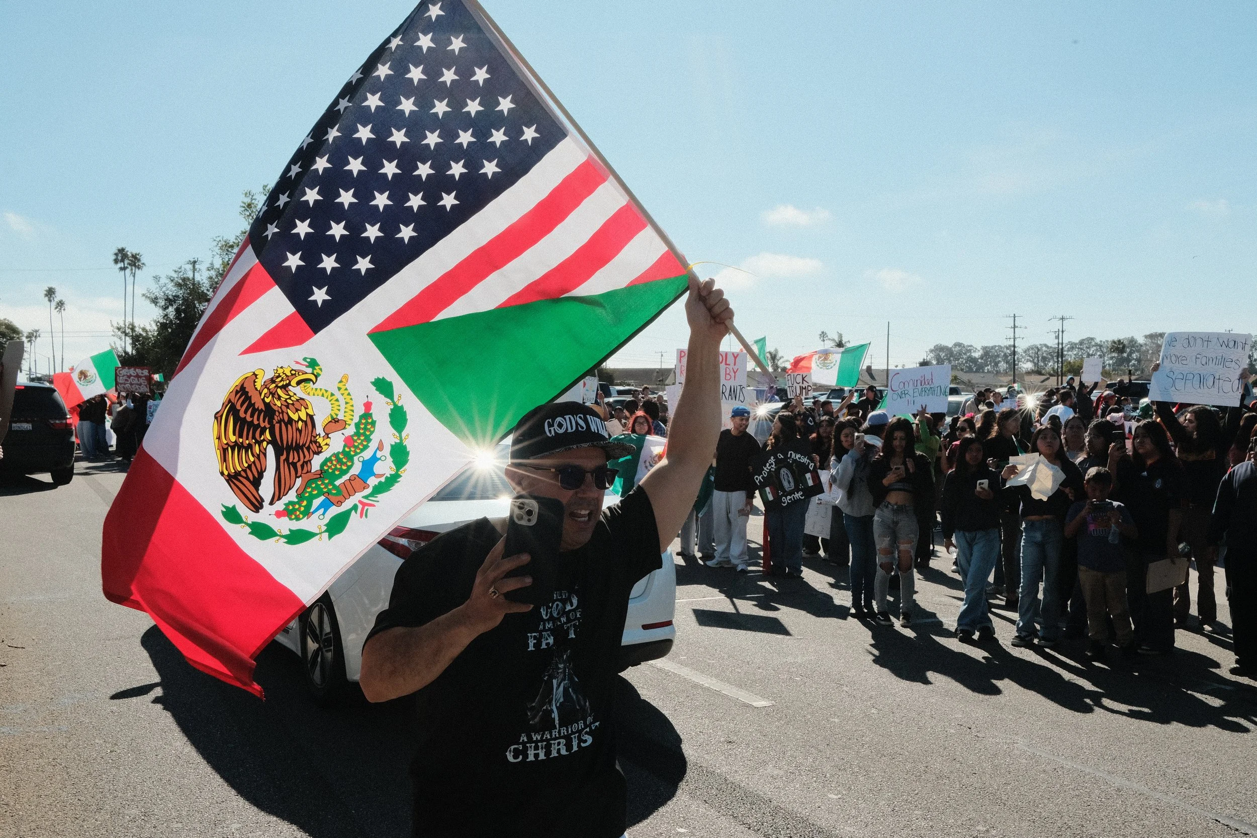 Person holding a large Mexican flag at a protest or rally, with a crowd holding signs in the background on a sunny day.