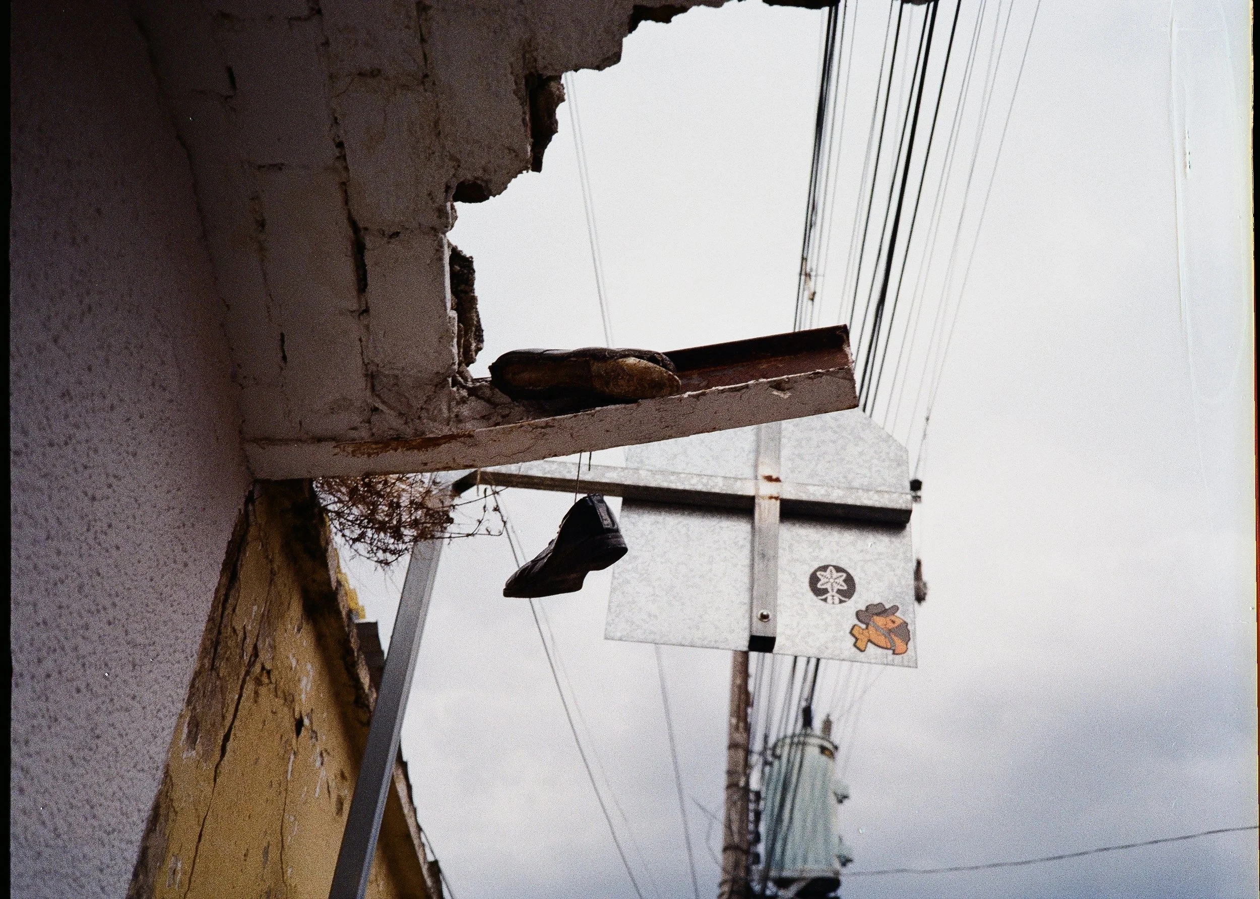 Close-up view of a weathered brick wall with a window opening, partially covered by a bracket or ledge with some objects on it. Visible power lines in the background and a gray cloudy sky.