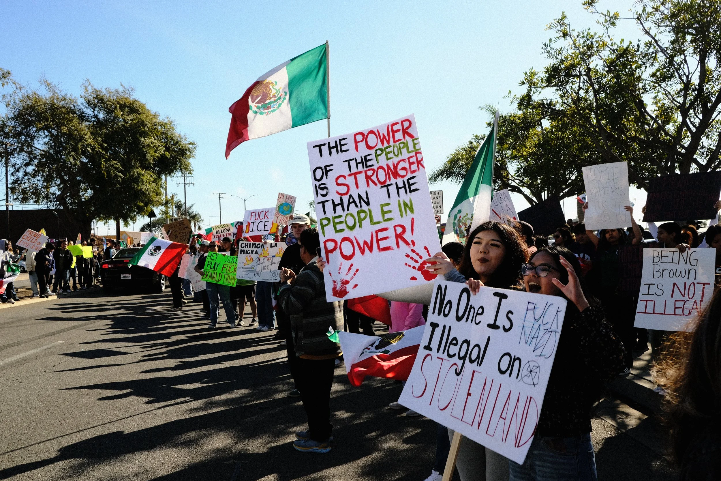 Protesters in a demonstration holding signs and flags, including a Mexican flag. Signs have messages advocating power for the people, criticizing immigration policies, and calling for equality and justice.