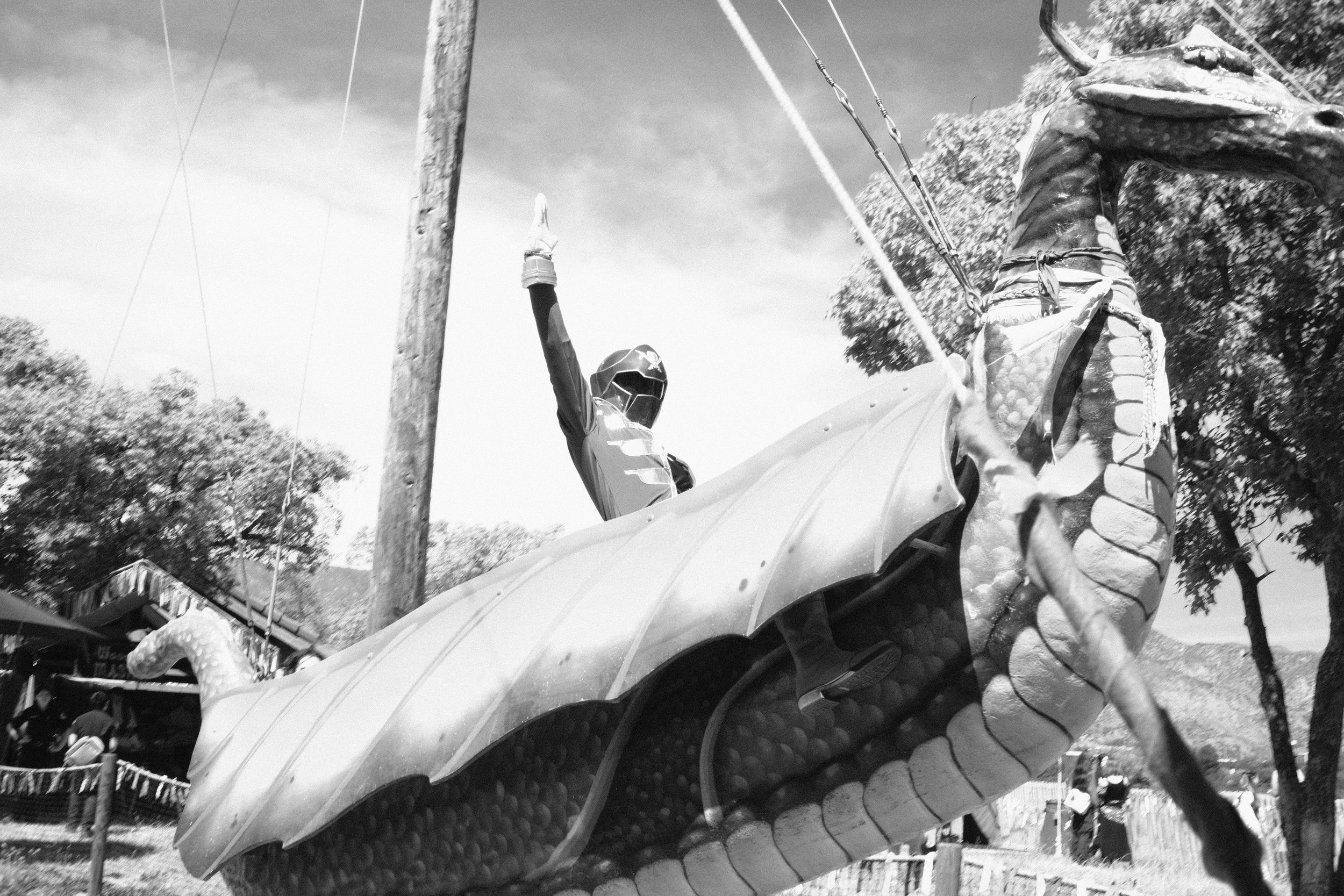 A drone with a dragon head design and wings flying over a carnival or fairground area with trees, tents, and people in the background.