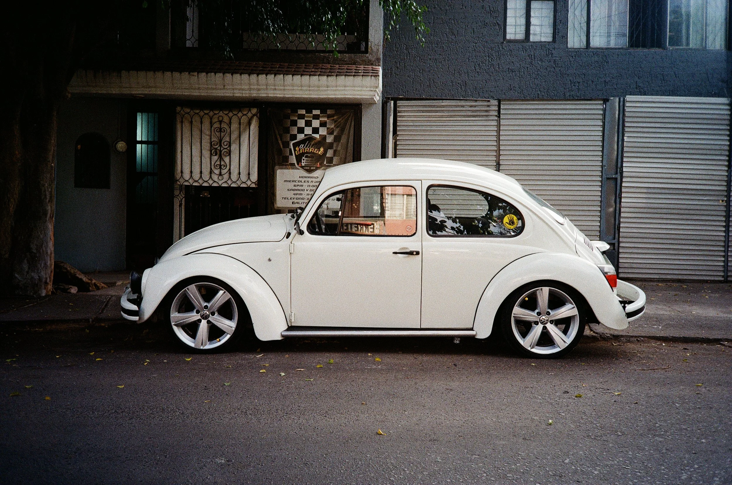 A white vintage Volkswagen Beetle parked on a street in front of a building with dark and light gray walls, a tree on the left, and a sign reading 'Auto Garage' behind the car.
