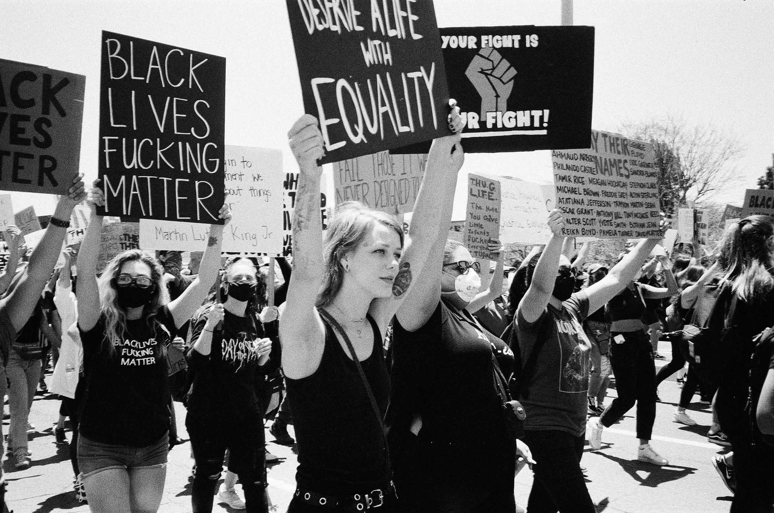 A group of protesters at a march, holding signs with messages supporting Black Lives Matter and social justice. Some signs read "Black Lives Fucking Matter," "Defund the Police with Equality," and "Your Fight is Our Fight." Many protesters are wearin