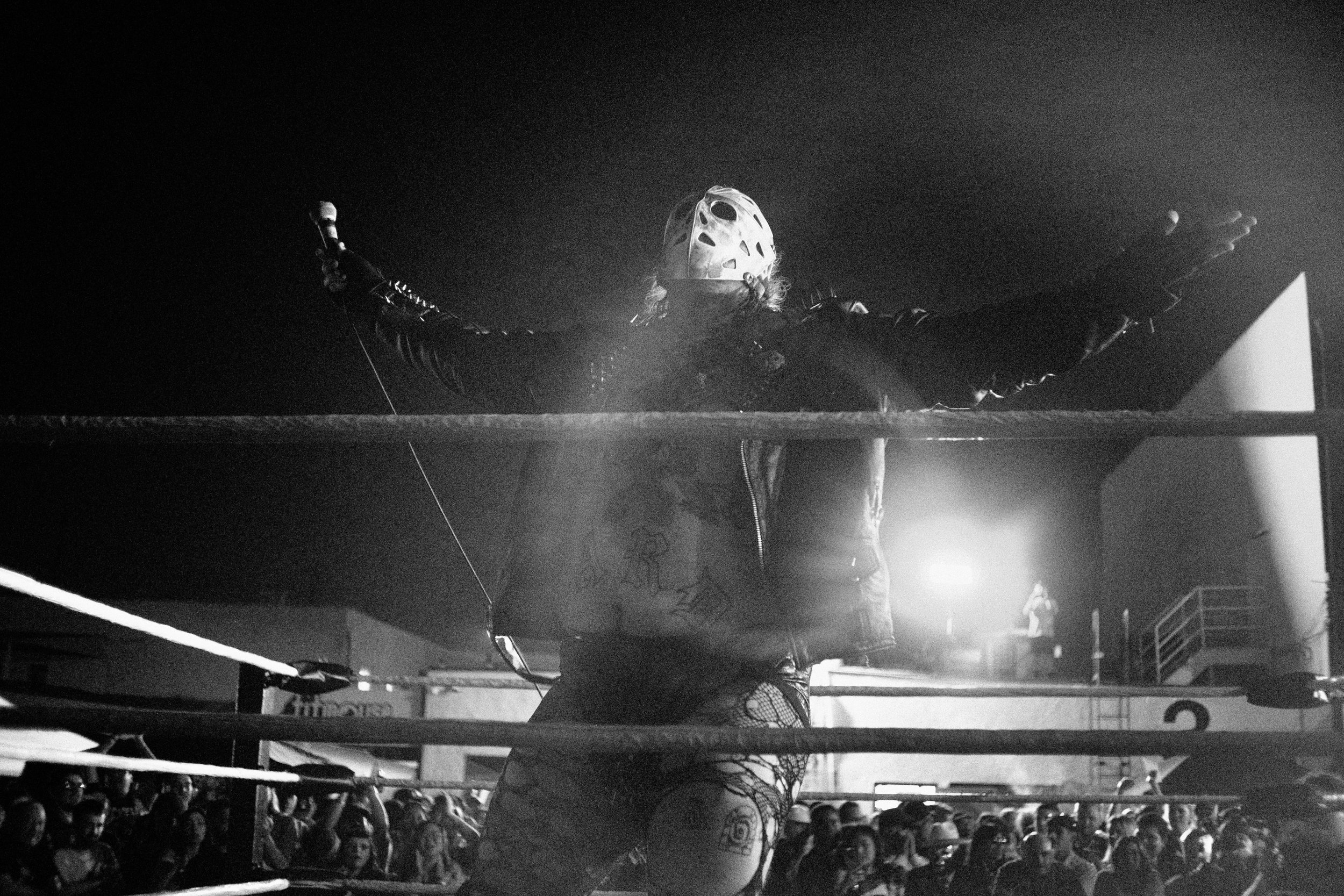 A person wearing a hockey mask and gloves, with tattoos visible on their arms and legs, standing in a wrestling ring with arms outstretched, while an audience watches below. The photo is black and white.