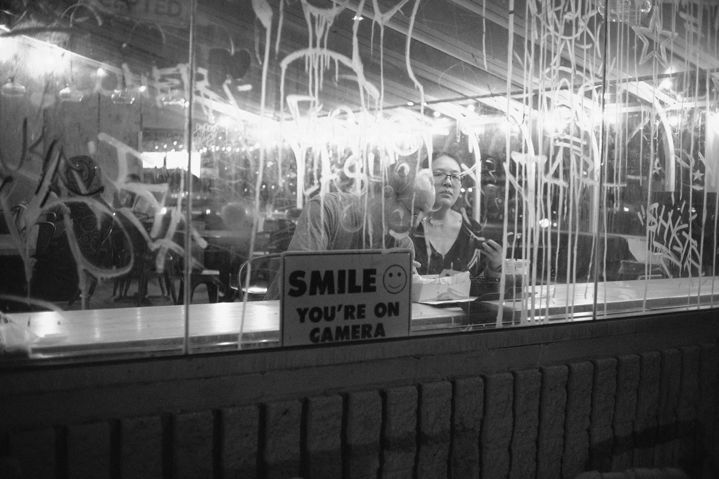 A person with glasses behind a glass window covered with graffiti, looking at the camera. There is a sign on the window that reads 'SMILE YOU'RE ON CAMERA'. The image is in black and white.