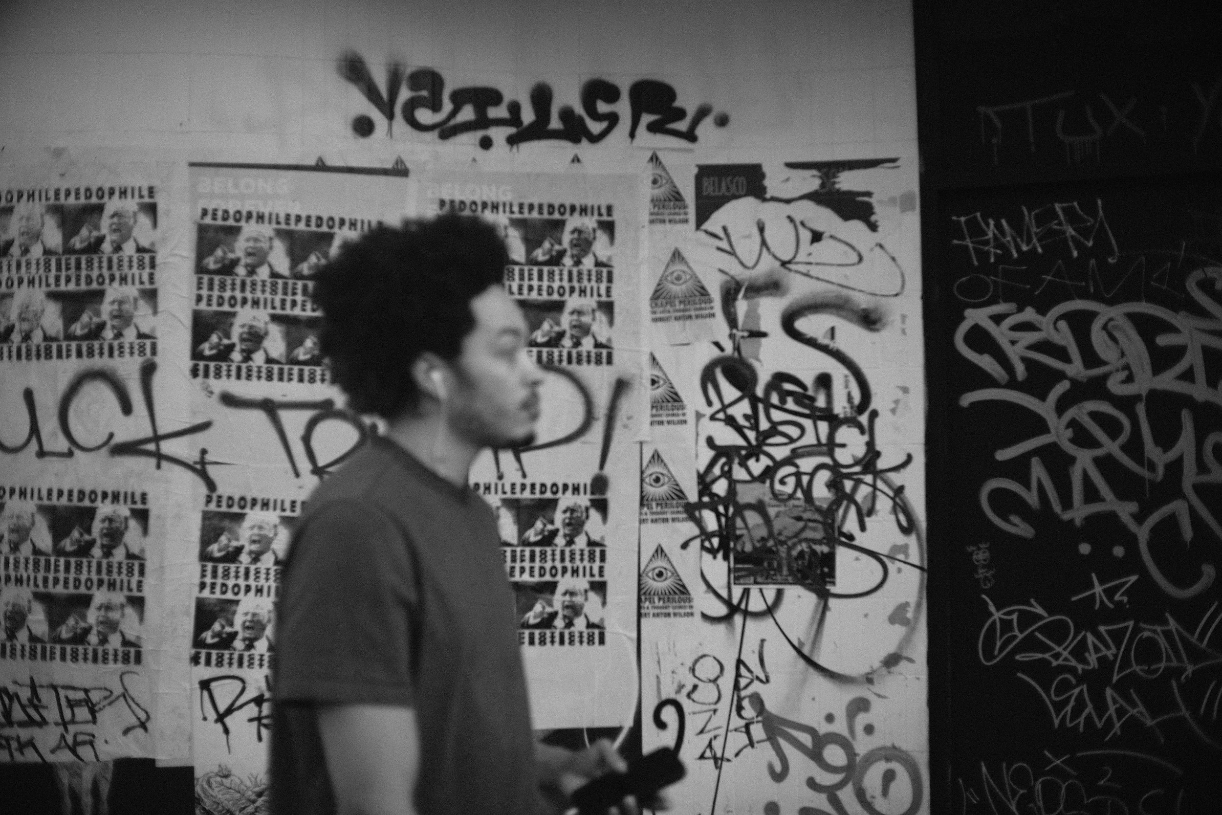 A person with curly dark hair walking past a wall covered with graffiti, posters, and stickers, including the word 'pedophile' and images of a talking person.