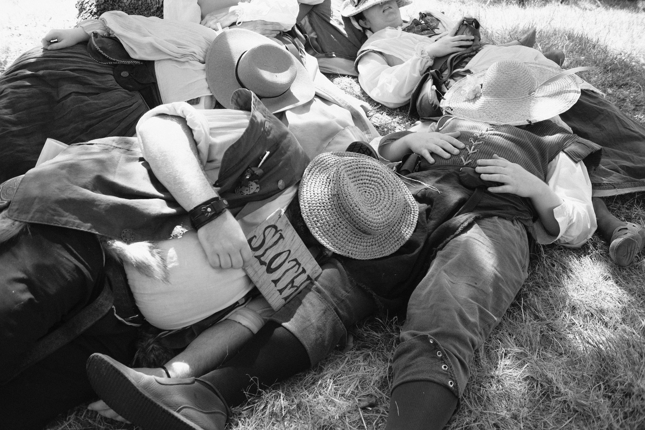 People lying down on the grass, wearing hats and casual outdoor clothing, some holding objects like a camera and a sign, in a park or open outdoor area.