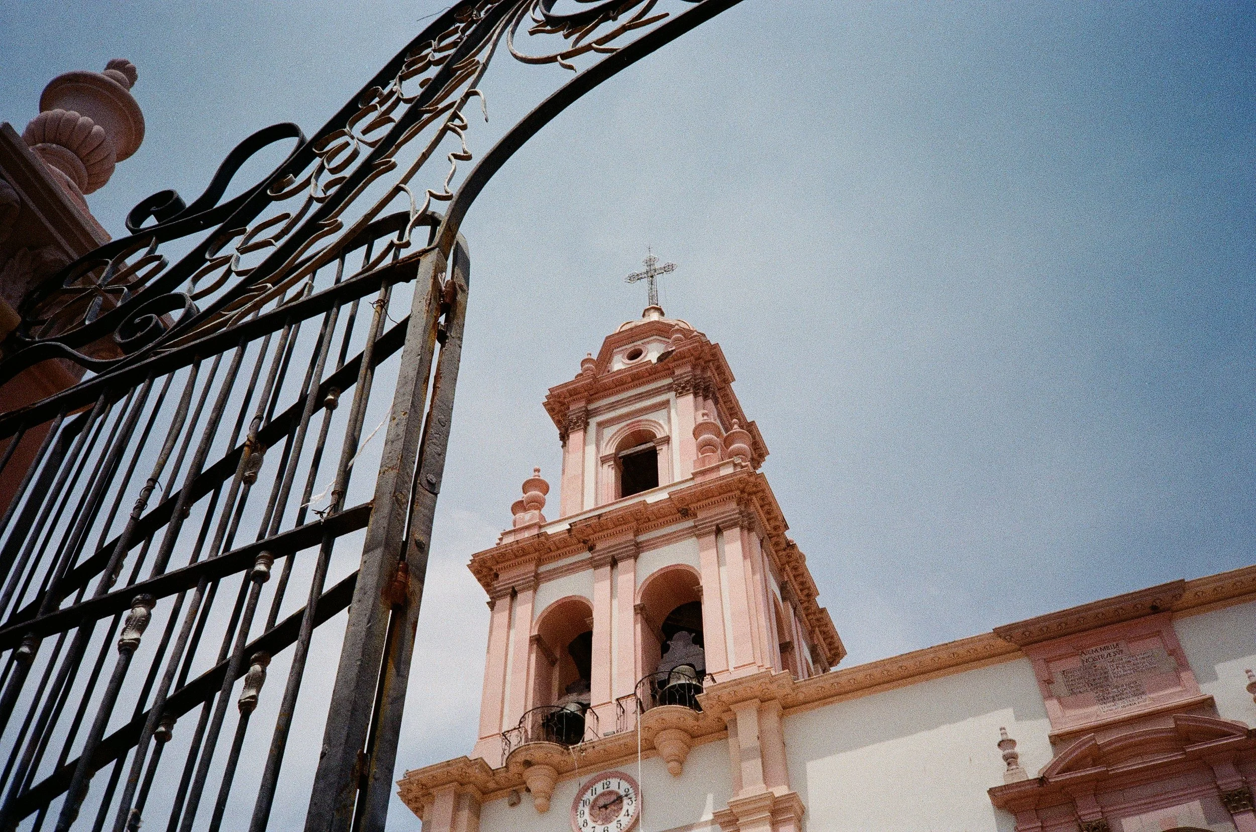 View of a historic church with a clock tower and a cross on top, behind an ornate wrought iron gate.