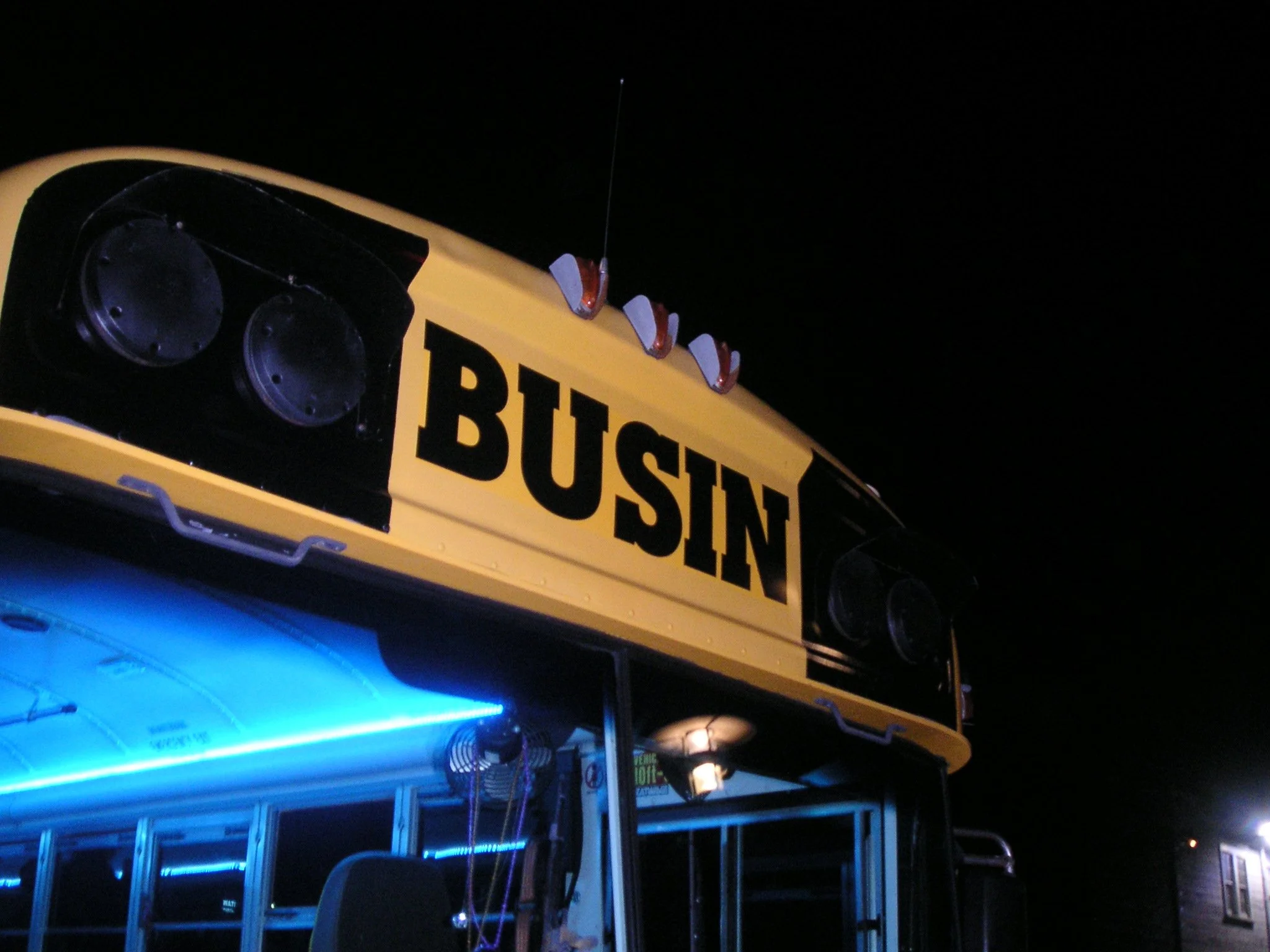 A yellow bus with the partial word 'BUSTIN' visible, decorated with orange and white lights on top, at night, illuminated by blue lights underneath, with a dark sky background.