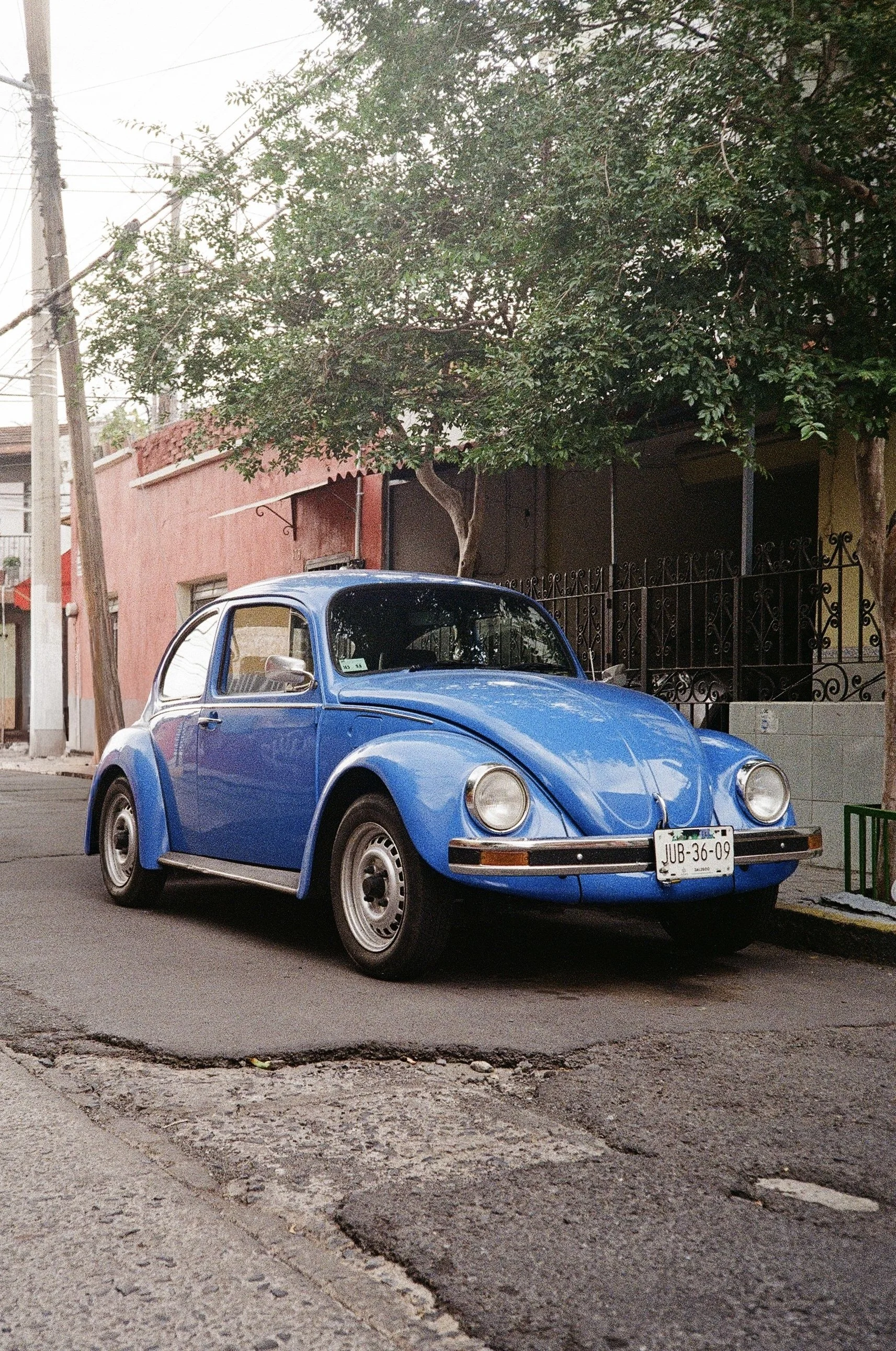 A blue vintage Volkswagen Beetle parked on a street in front of a pink and yellow building with a tree and a black wrought iron fence.