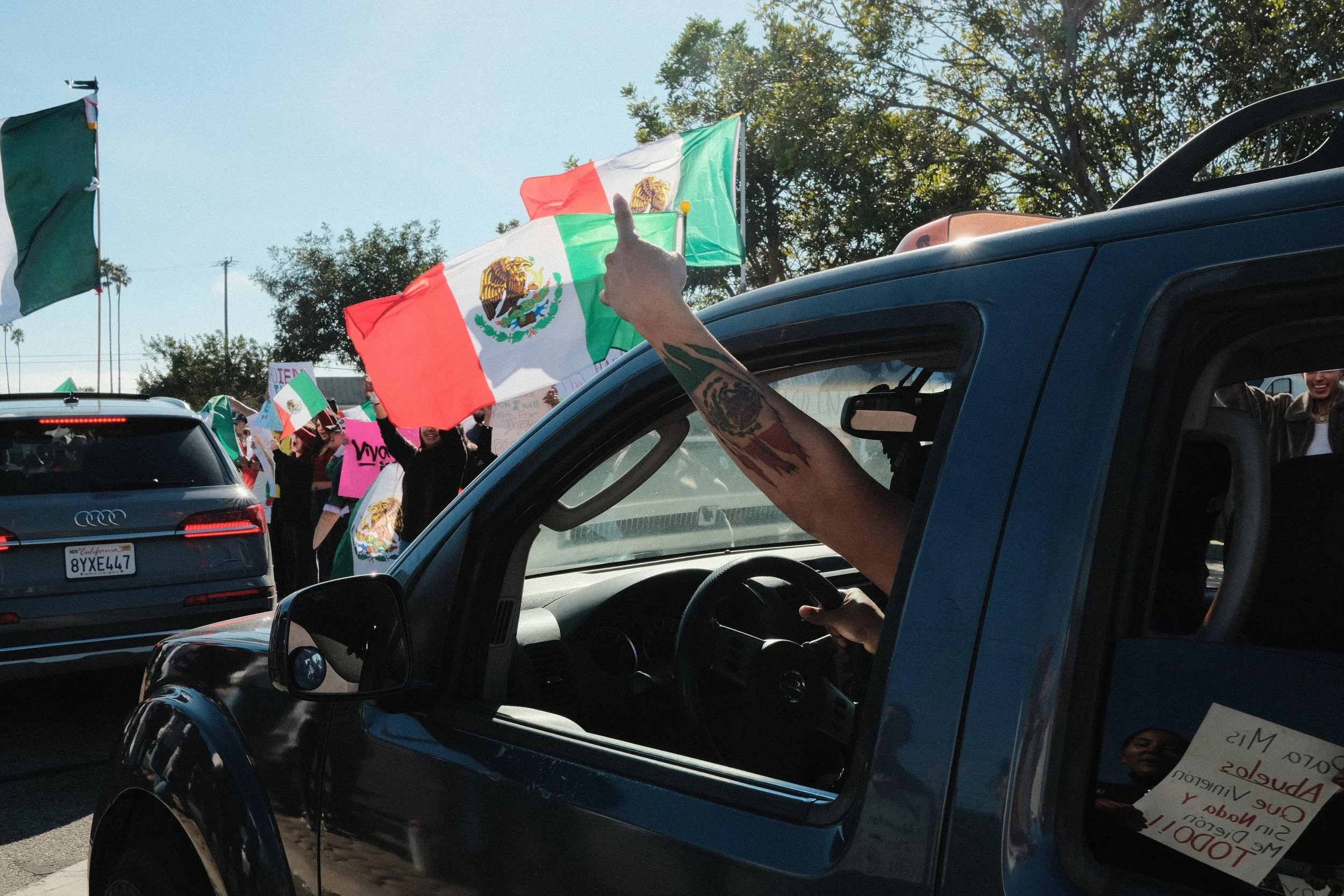 People holding Mexican flags and protest signs during a demonstration near cars on a street.