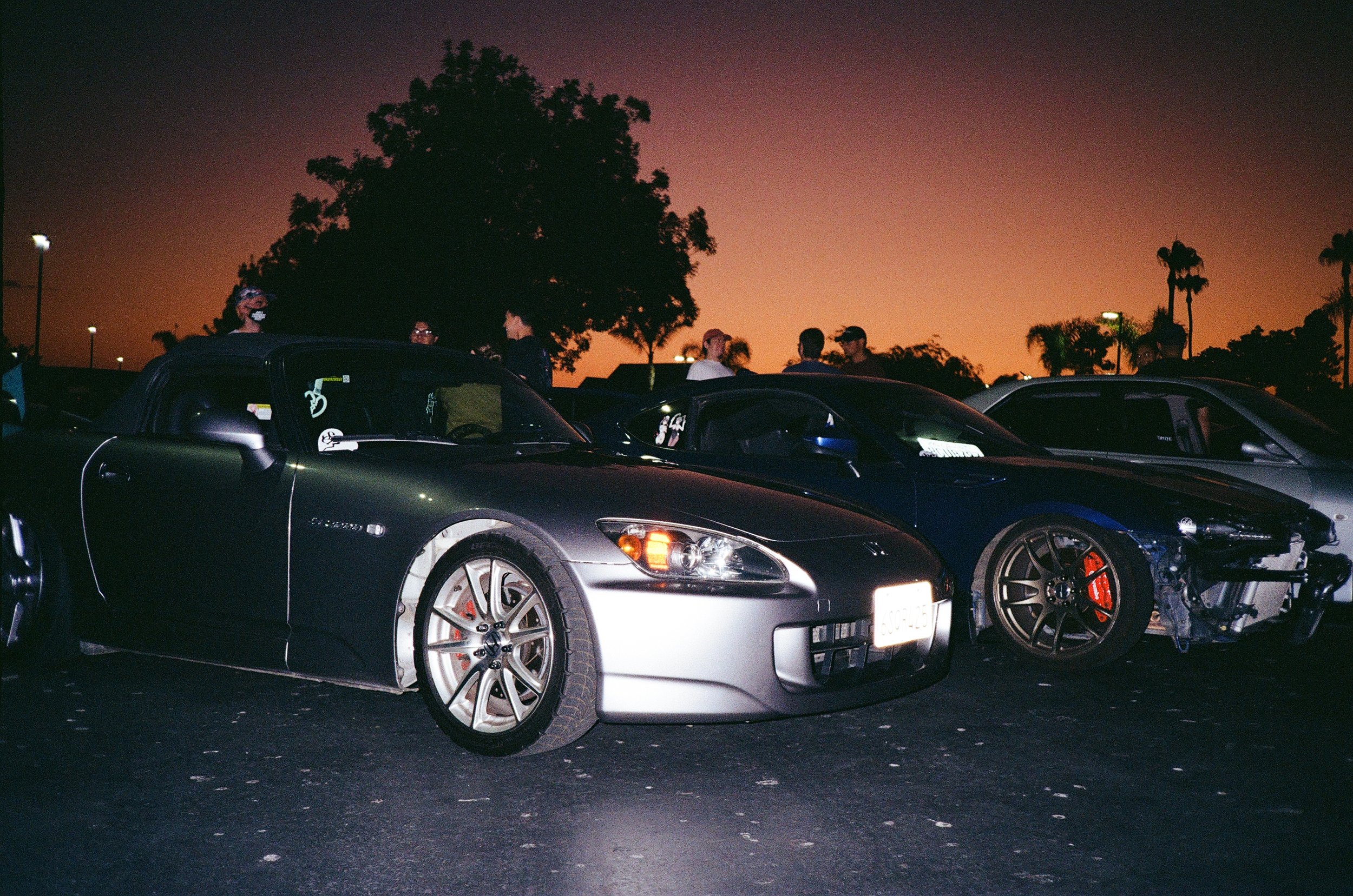 A group of people gathered around parked cars in a parking lot at dusk, with silhouettes of trees and a colorful sky in the background.