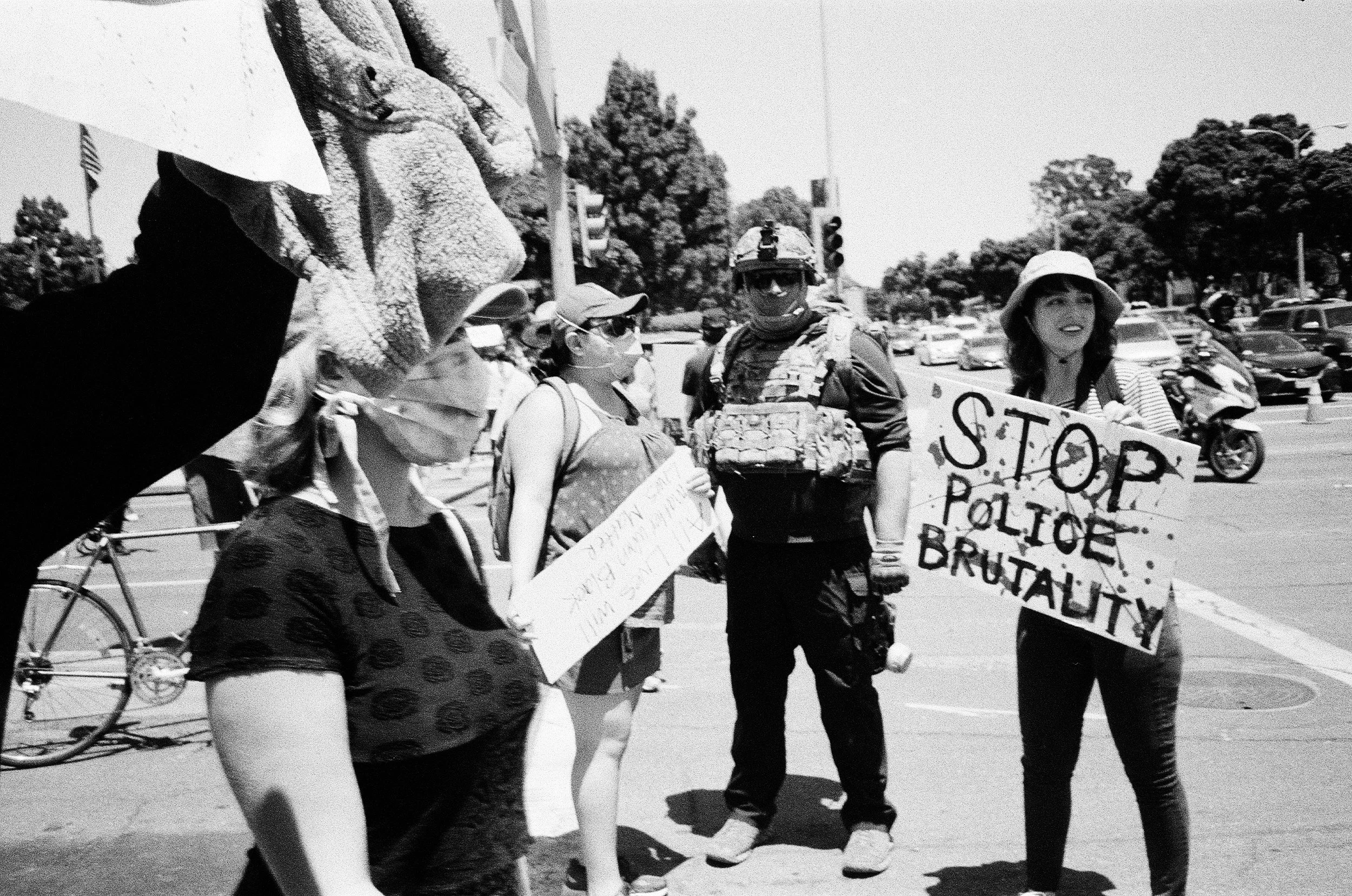 Protesters holding signs during a demonstration, with a military officer present, on a city street with trees and traffic in the background. One sign reads 'STOP POLICE BRUTALITY'.