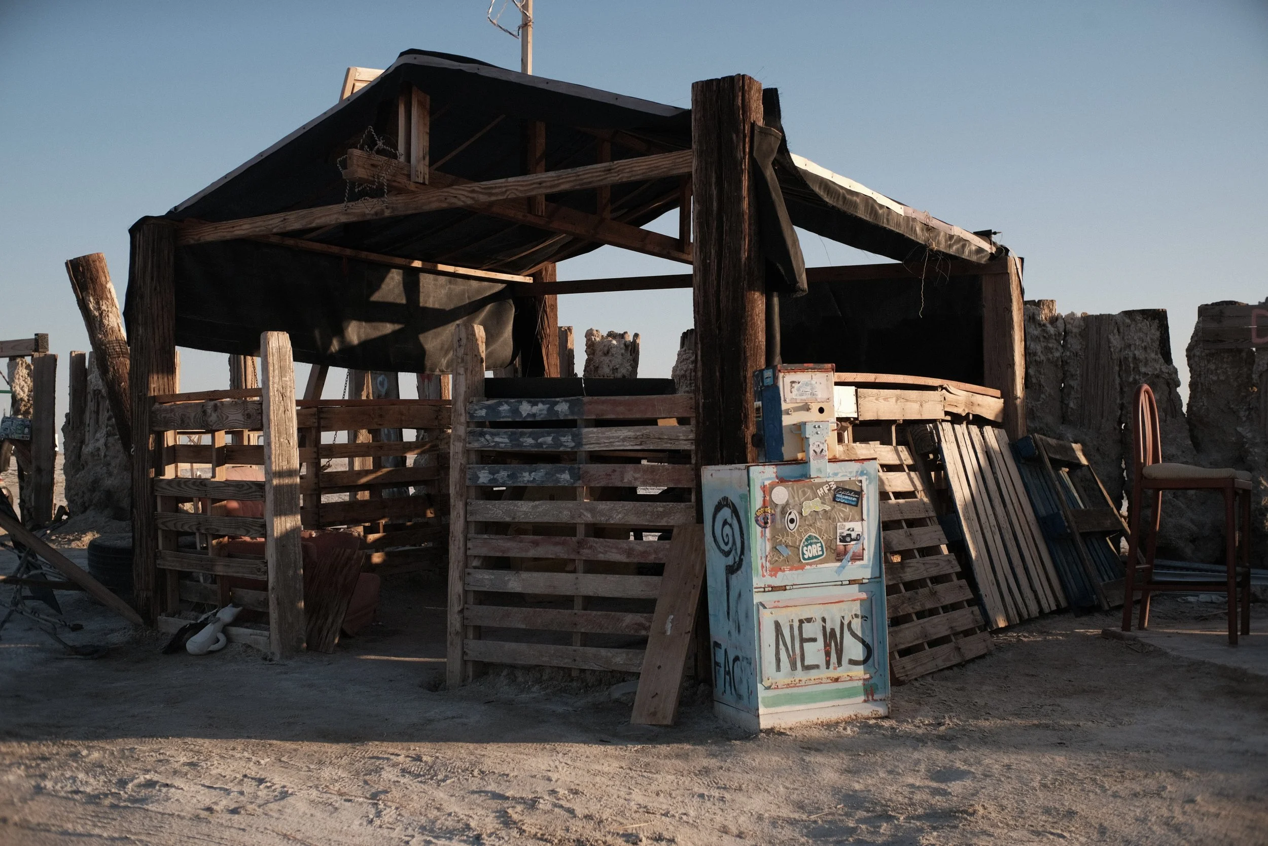 A makeshift structure built from wooden pallets and scrap materials with a sign that reads 'NEWS' on it. The structure has a roof made of wooden planks and is situated outdoors under a clear blue sky.