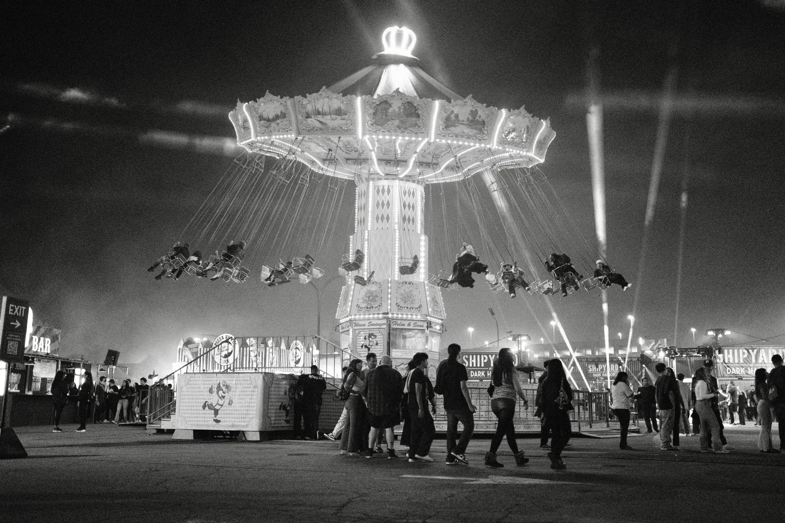 A black and white photo of a carousel ride at night with people standing and walking nearby. The carousel is lit up with bright lights and has a crown ornament on top. There are signs for food and drink at the bottom of the image.